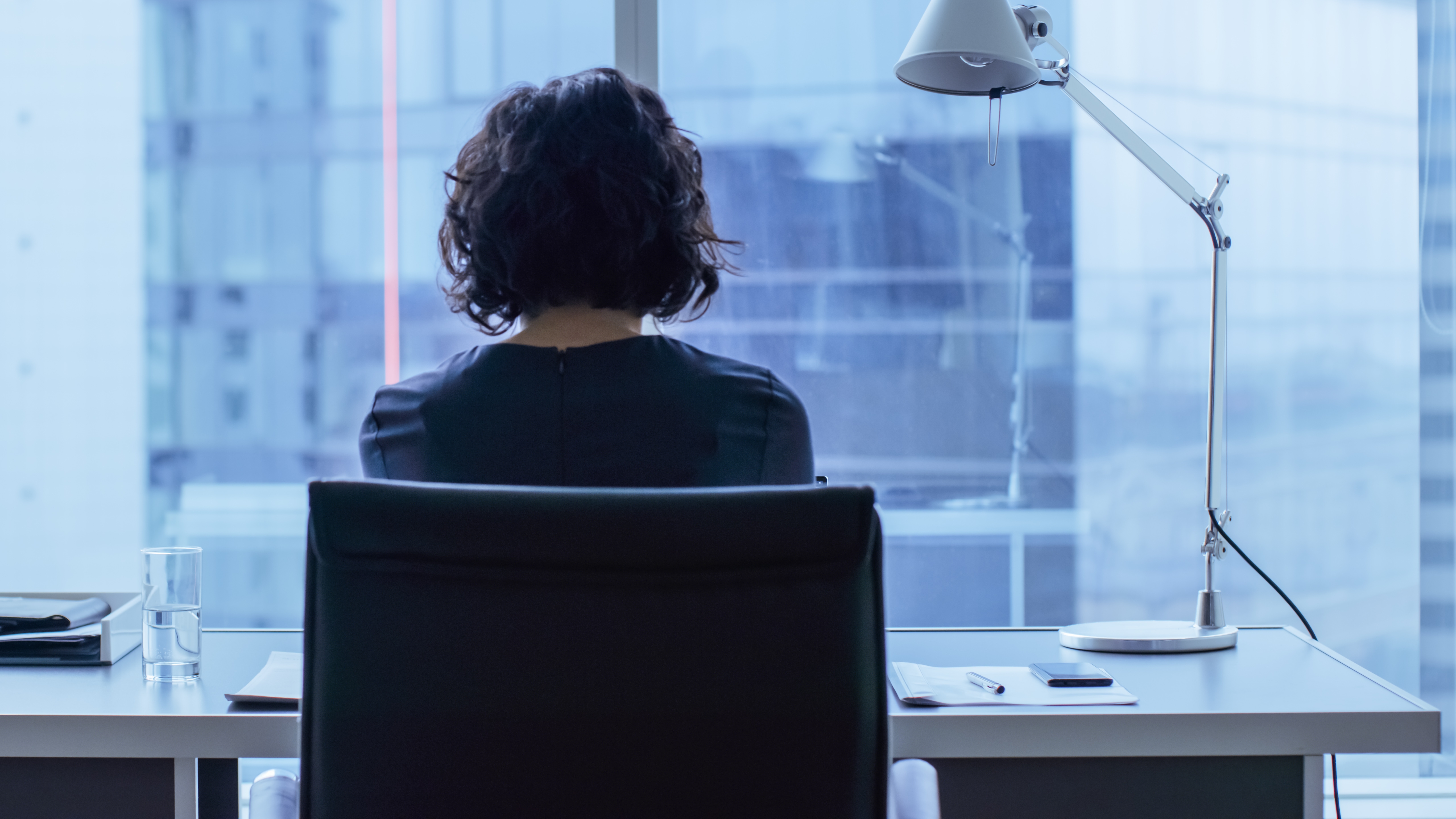 Person med kort hår sitter ved et skrivebord på et kontor og vender mot et stort vindu. Skrivebordet holder en lampe, vannglass og papirer. Scenen er rolig og introspektiv