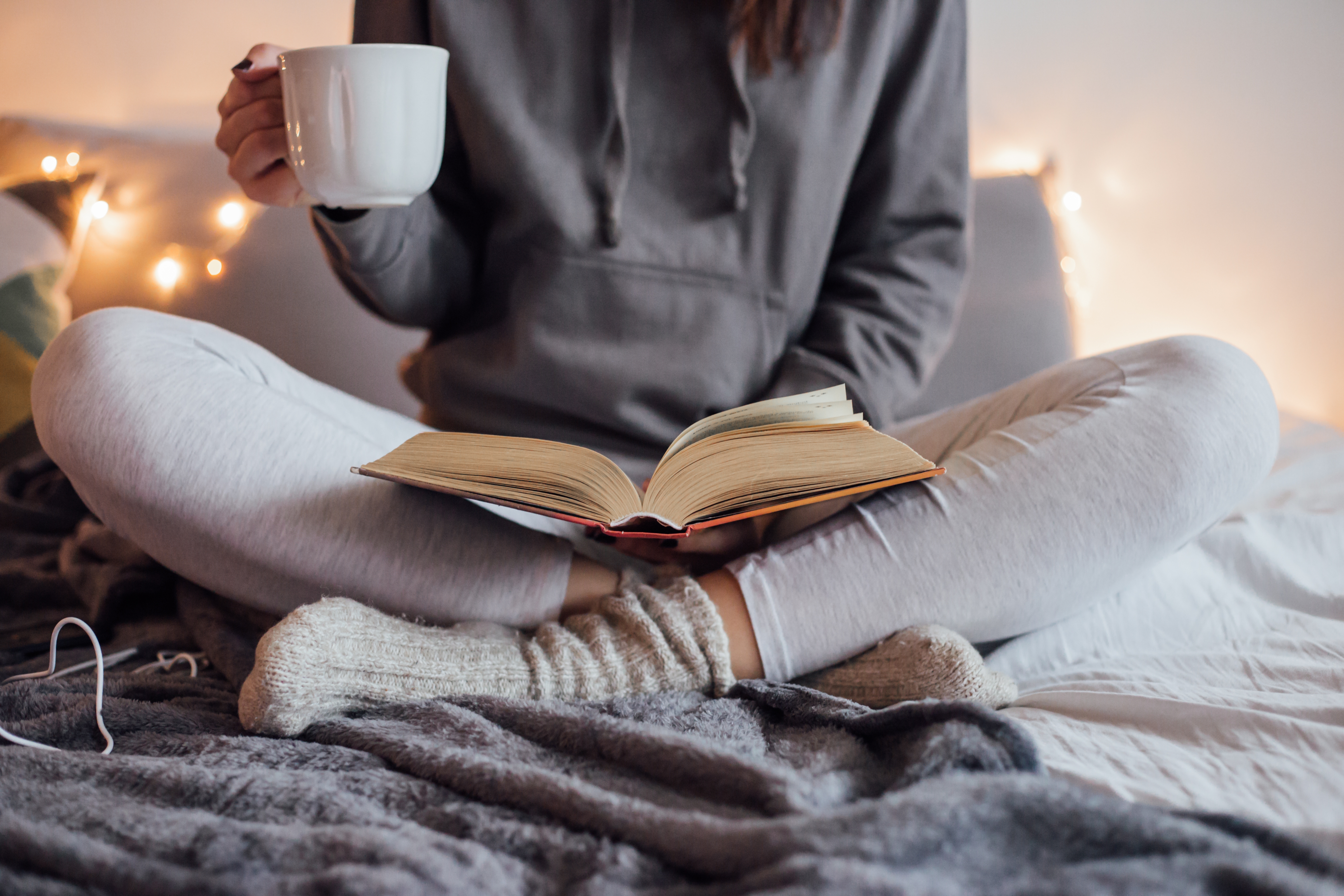 Person sitting cross-legged on a bed, wearing a hoodie and socks, holding a mug and reading a book, surrounded by cozy blankets and string lights