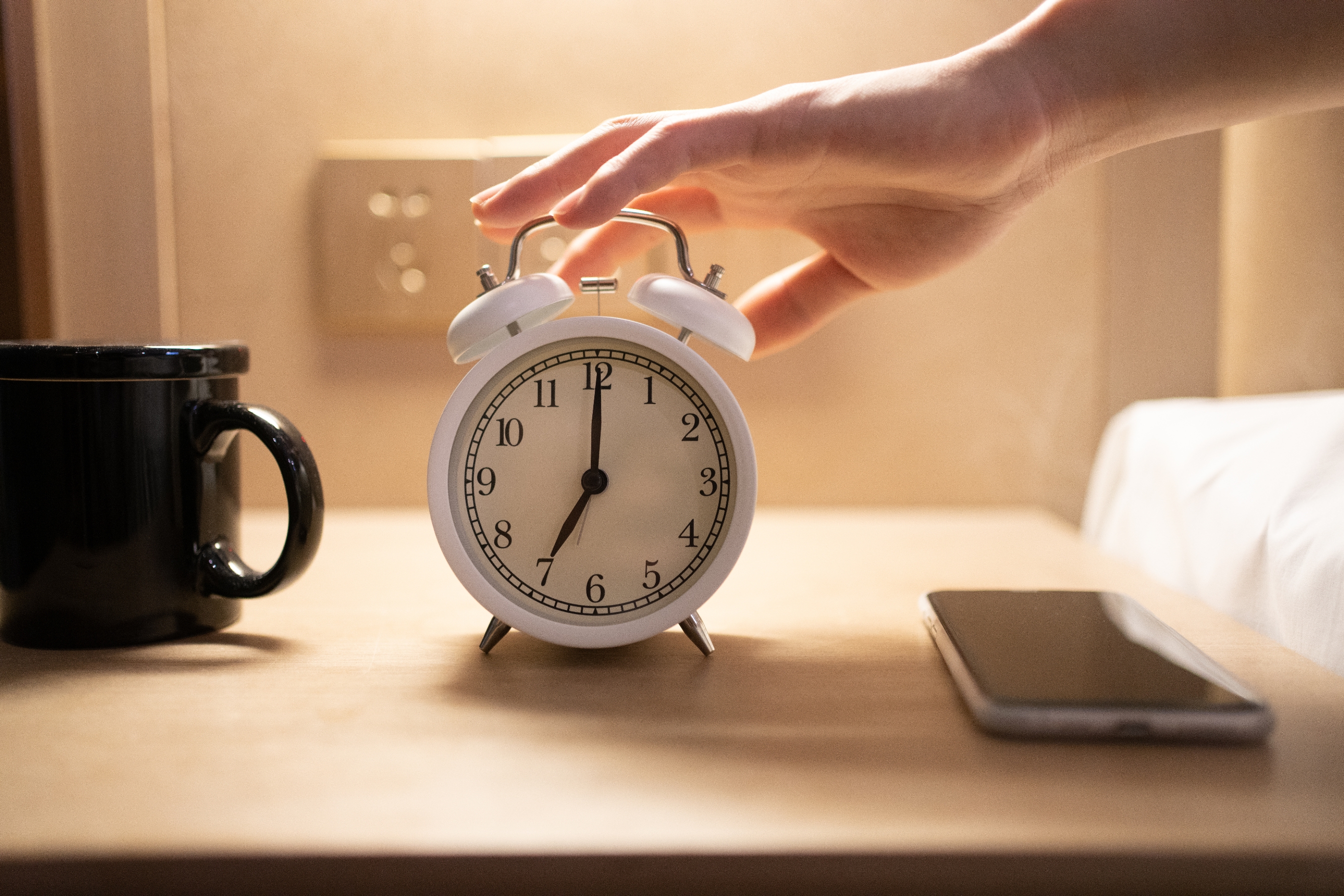 A hand reaches to turn off a ringing alarm clock on a bedside table, next to a smartphone and mug
