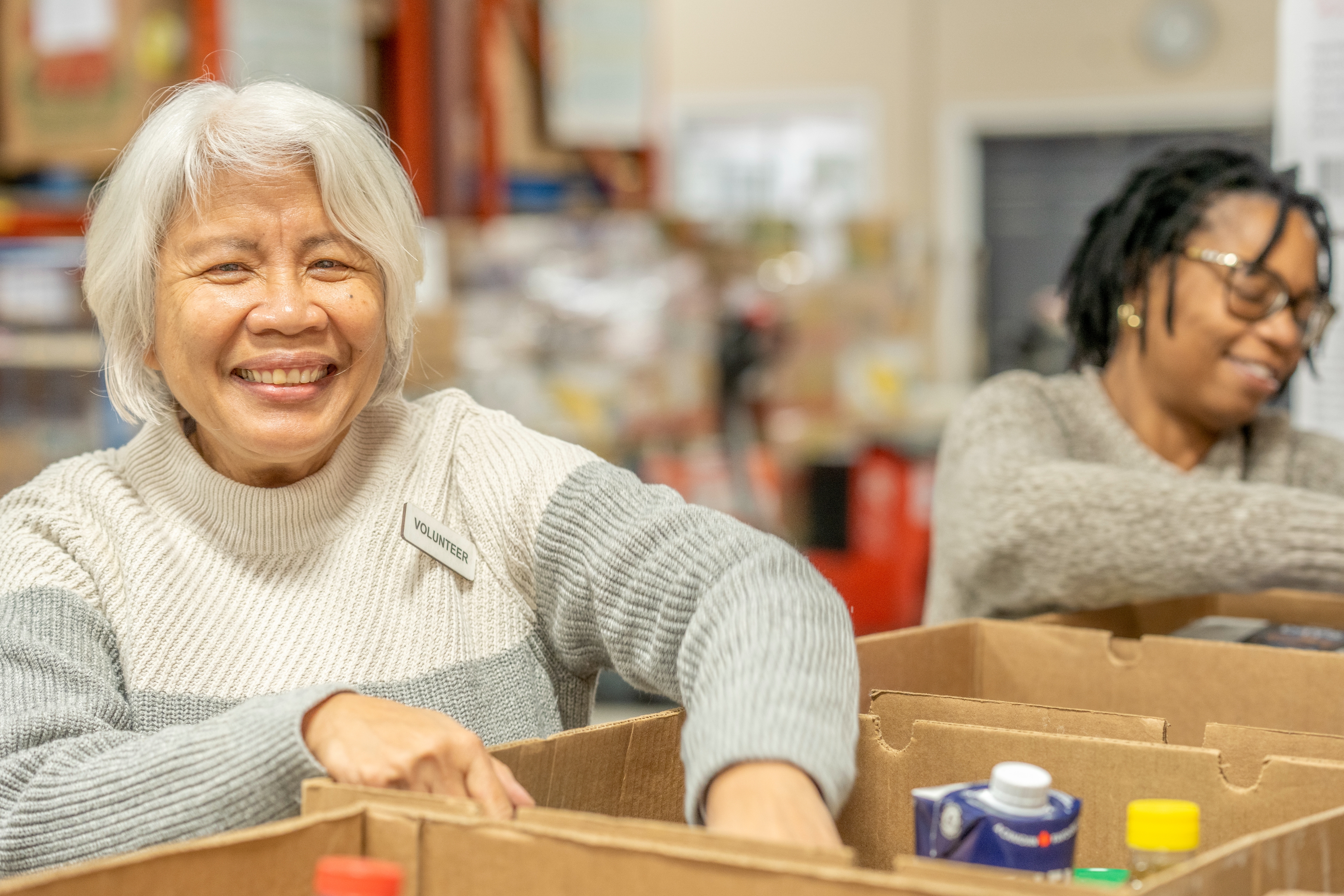 Two people happily pack boxes with food items in a busy community center