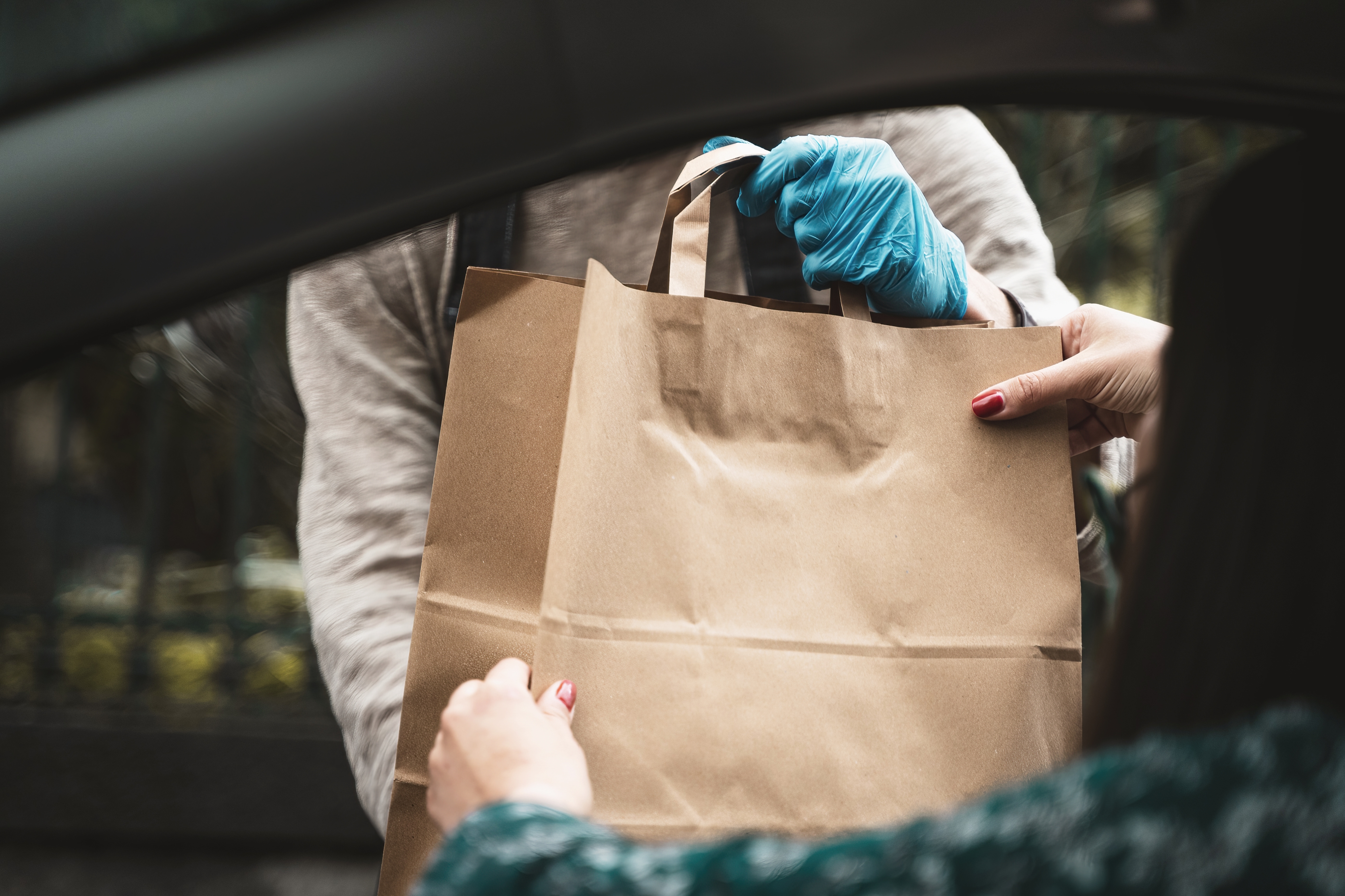 Person in gloves hands a brown paper bag to someone through a car window, suggesting a contactless food or package delivery