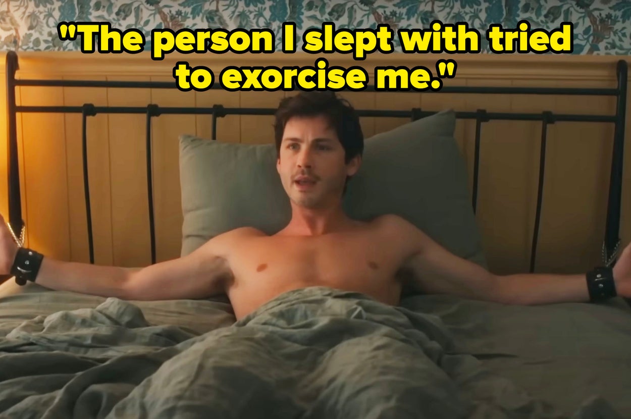 Shirtless man with outstretched arms and wristbands, sitting up in bed with patterned wallpaper in the background