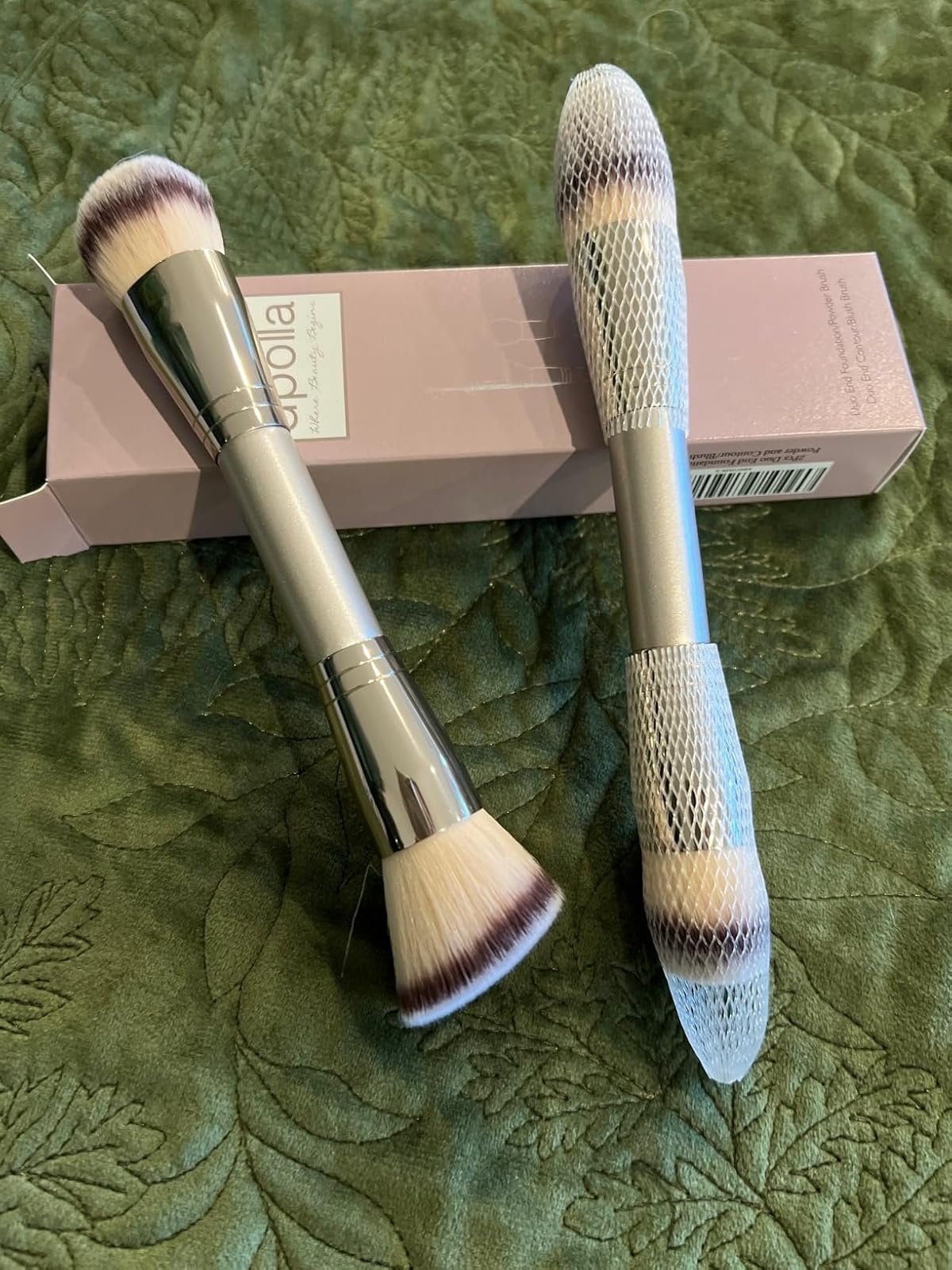 Two double-ended makeup brushes with protective nets displayed on a green surface, next to a branded box