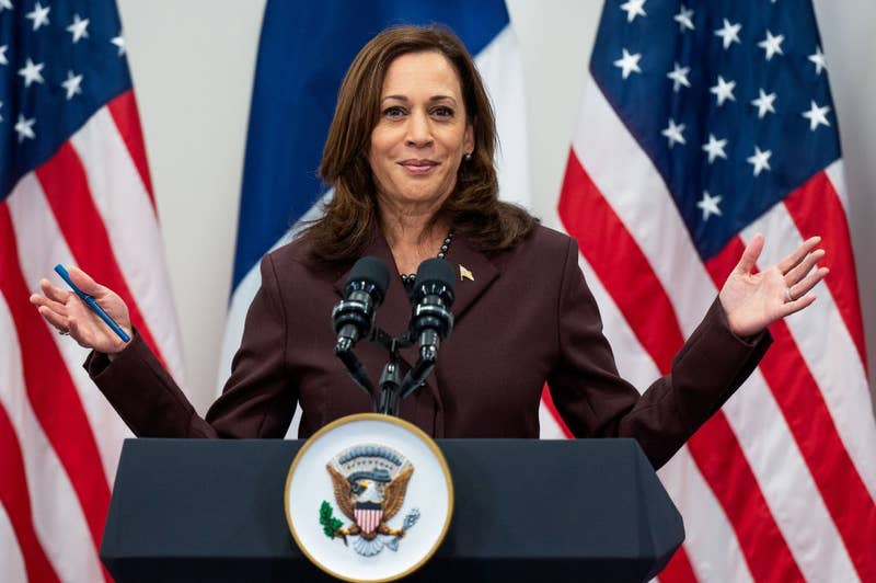 A public figure speaks at a podium with U.S. flags in the background, wearing a formal suit, gesturing with both hands open