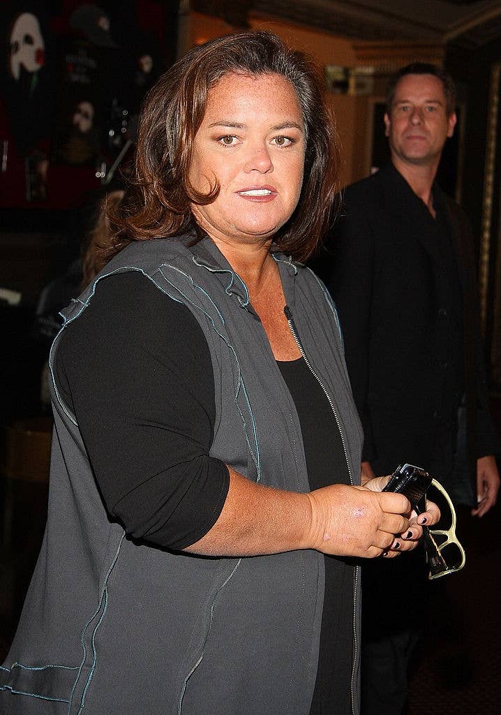Person in a casual black and gray outfit holding sunglasses, standing indoors at an event. Another person is in the background