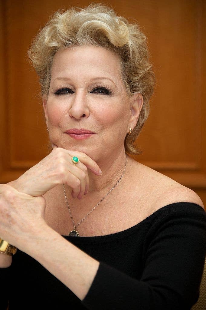 Person with short, styled hair in an off-the-shoulder top, resting their chin on their hand, wearing a ring and bracelet