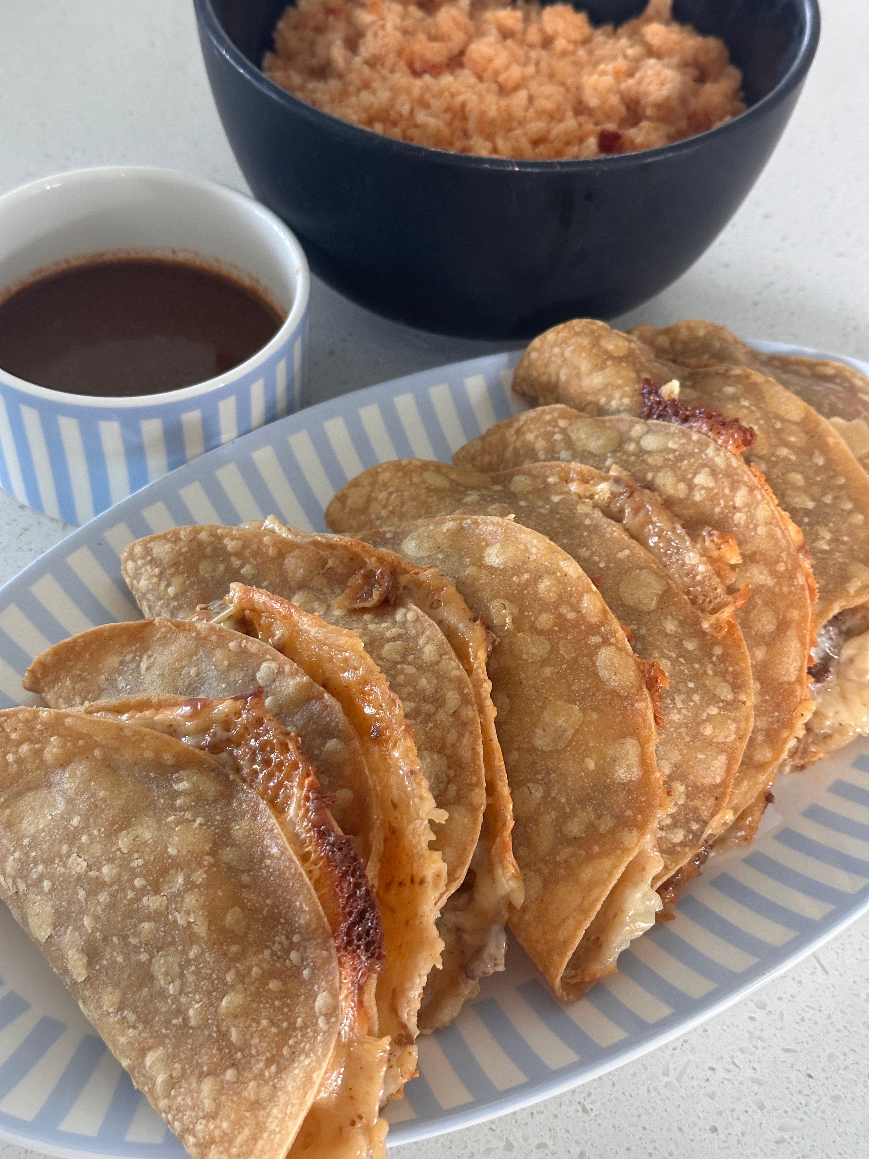 Plate of folded tacos with cheese and meat, beside a cup of sauce and a bowl of rice