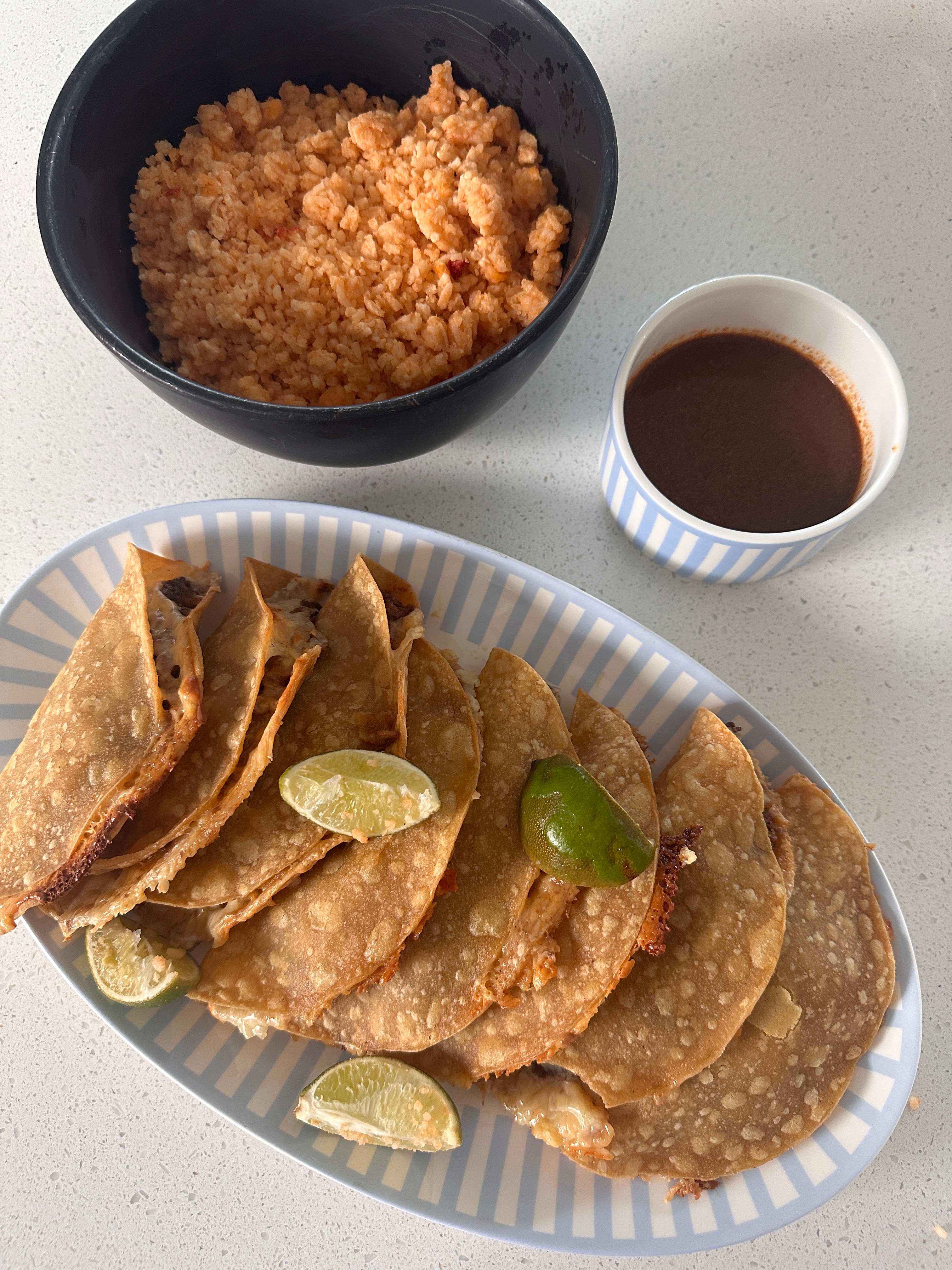 Tacos with lime wedges laid on a plate next to a bowl of rice and a cup of sauce on a countertop