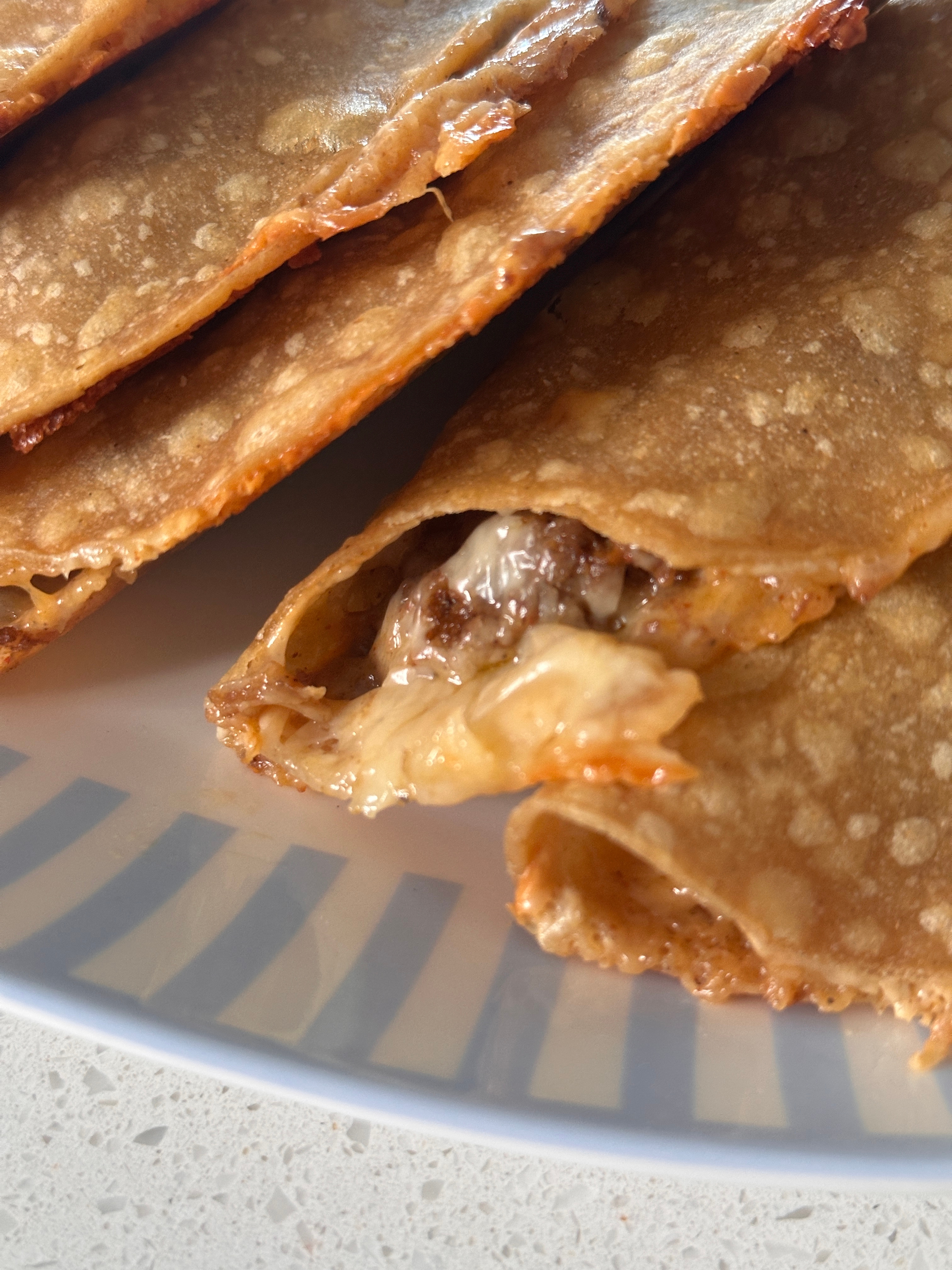 Close-up of folded, crispy tacos filled with melted cheese and beef on a plate with a striped pattern