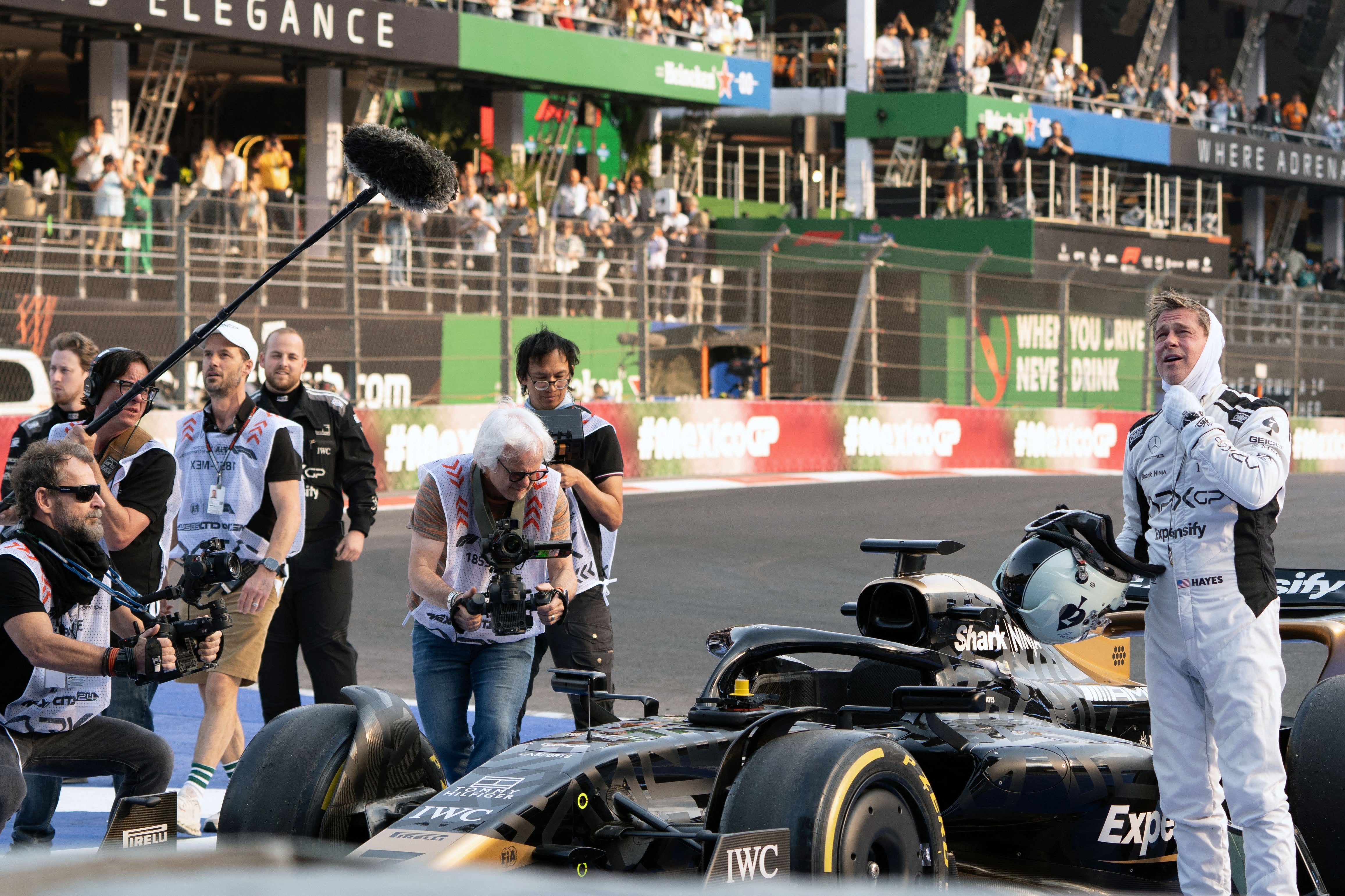 Racecar driver in a racing suit enthusiastically interacts with film crew beside a parked F1 car on the track, with cameras and boom mic visible