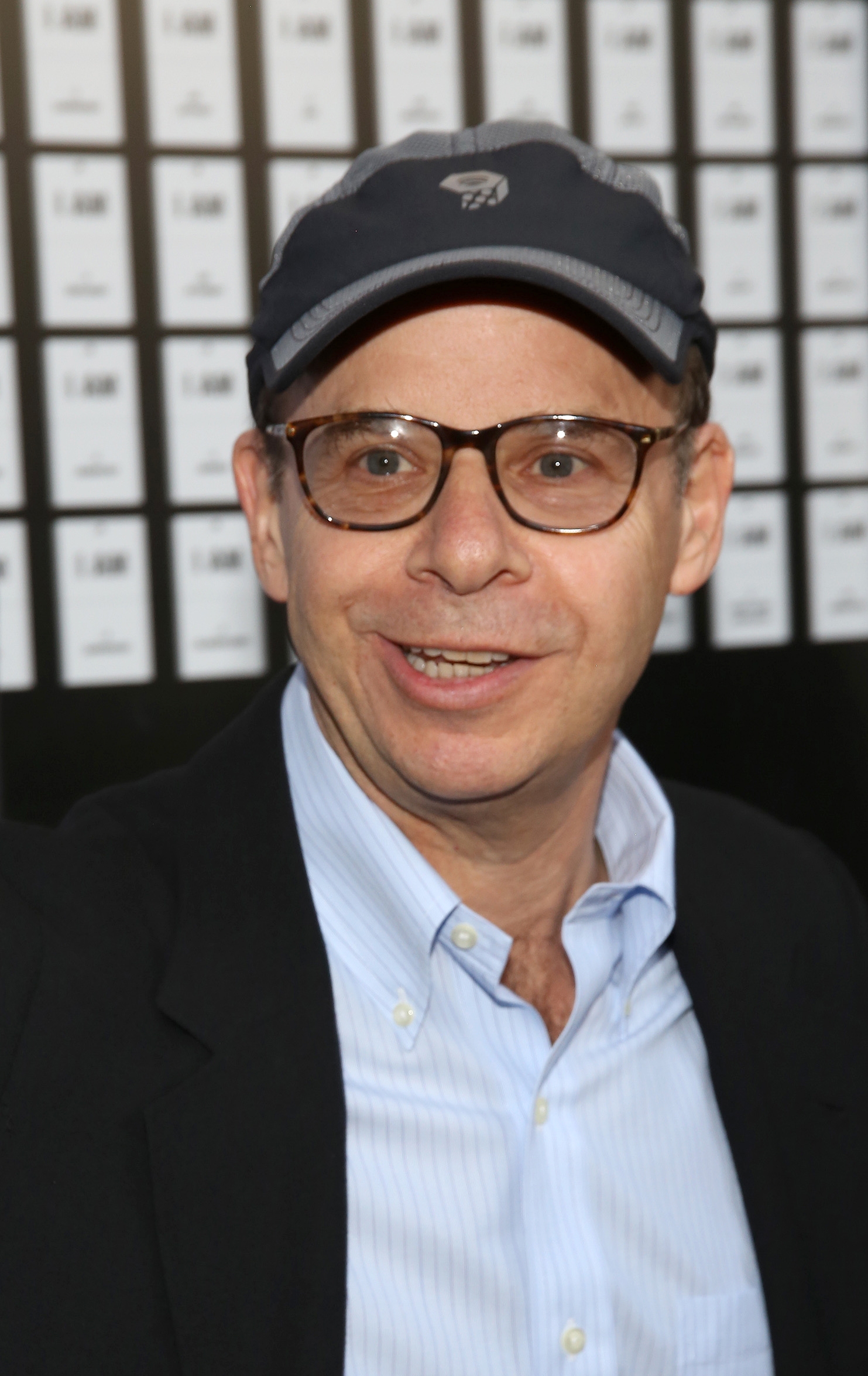 Person wearing glasses, baseball cap, and open-collar shirt attends event with background featuring repeating text