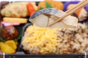 割り箸で持ち上げられた焼き魚が中央にあり、背景に盛り付けられた様々な和食のお弁当が見えます。