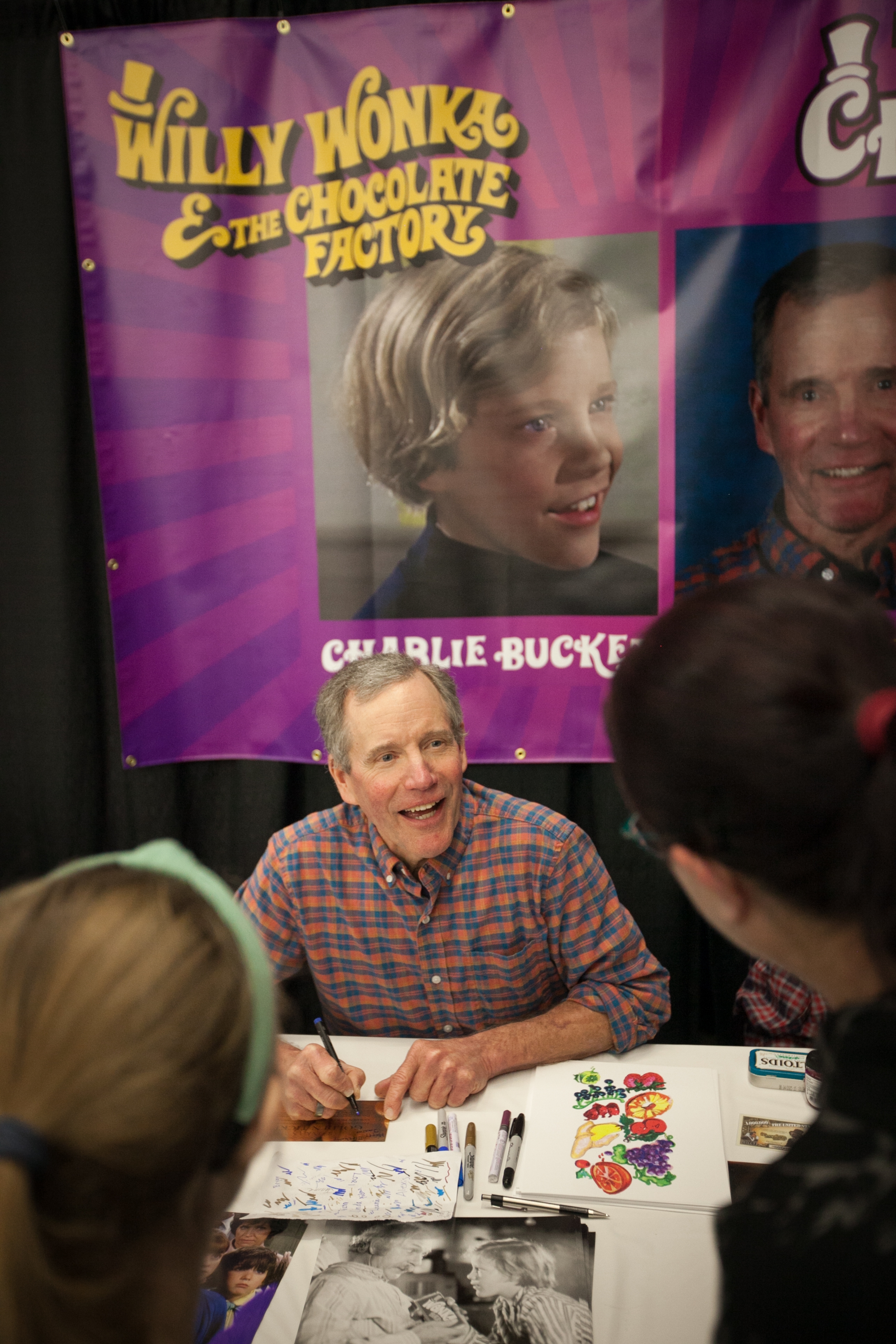 Person signing autographs at an event, sitting at a table with "Willy Wonka &amp;amp; the Chocolate Factory" posters