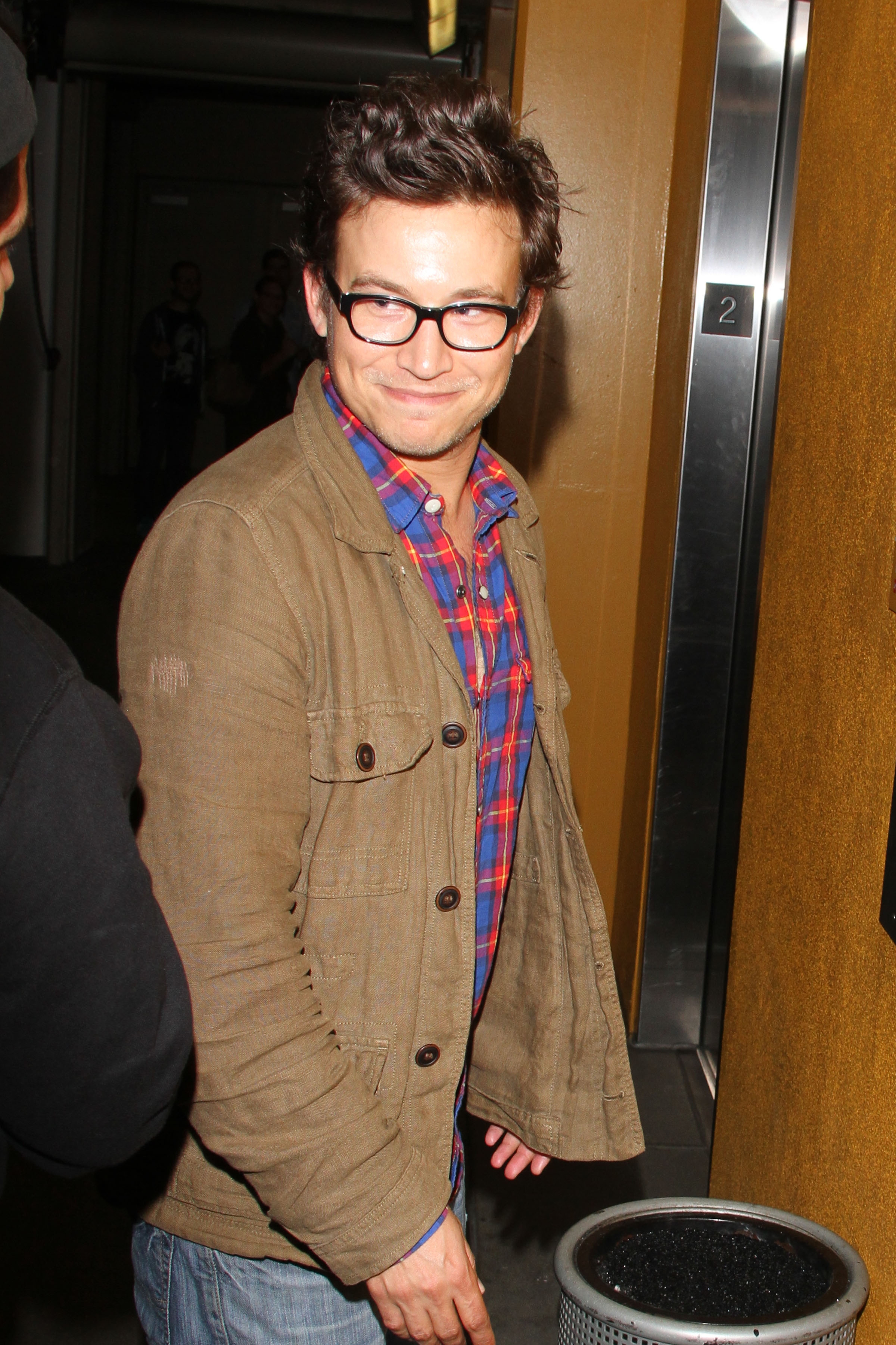 A person in glasses wears a casual brown jacket over a plaid shirt, smiling while walking indoors. An elevator and another person are in the background