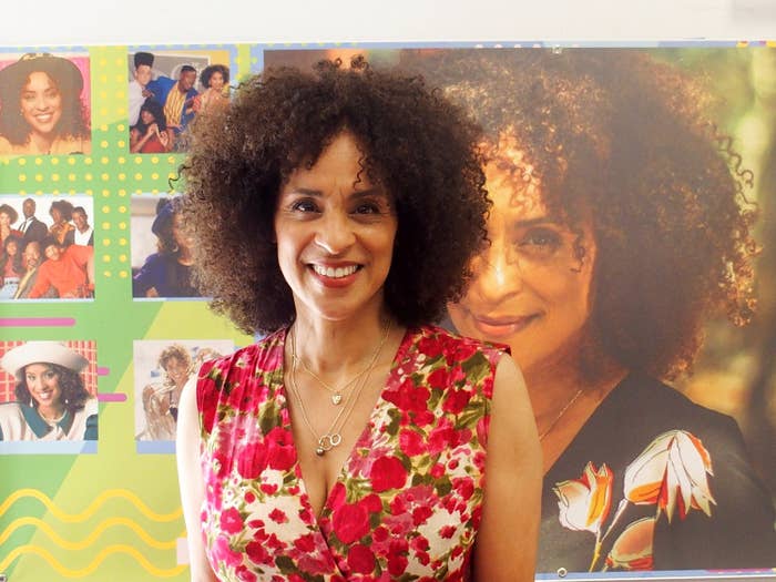Woman with curly hair in floral dress stands smiling in front of a collage of photos from various periods