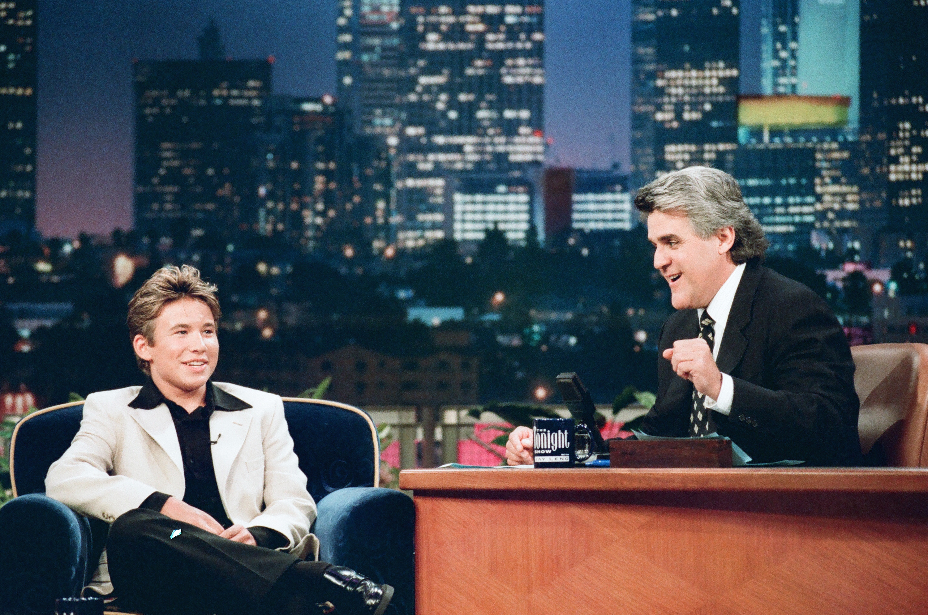 Talk show host interviewing a guest on a late-night set with a cityscape backdrop; both are smiling and dressed in formal attire