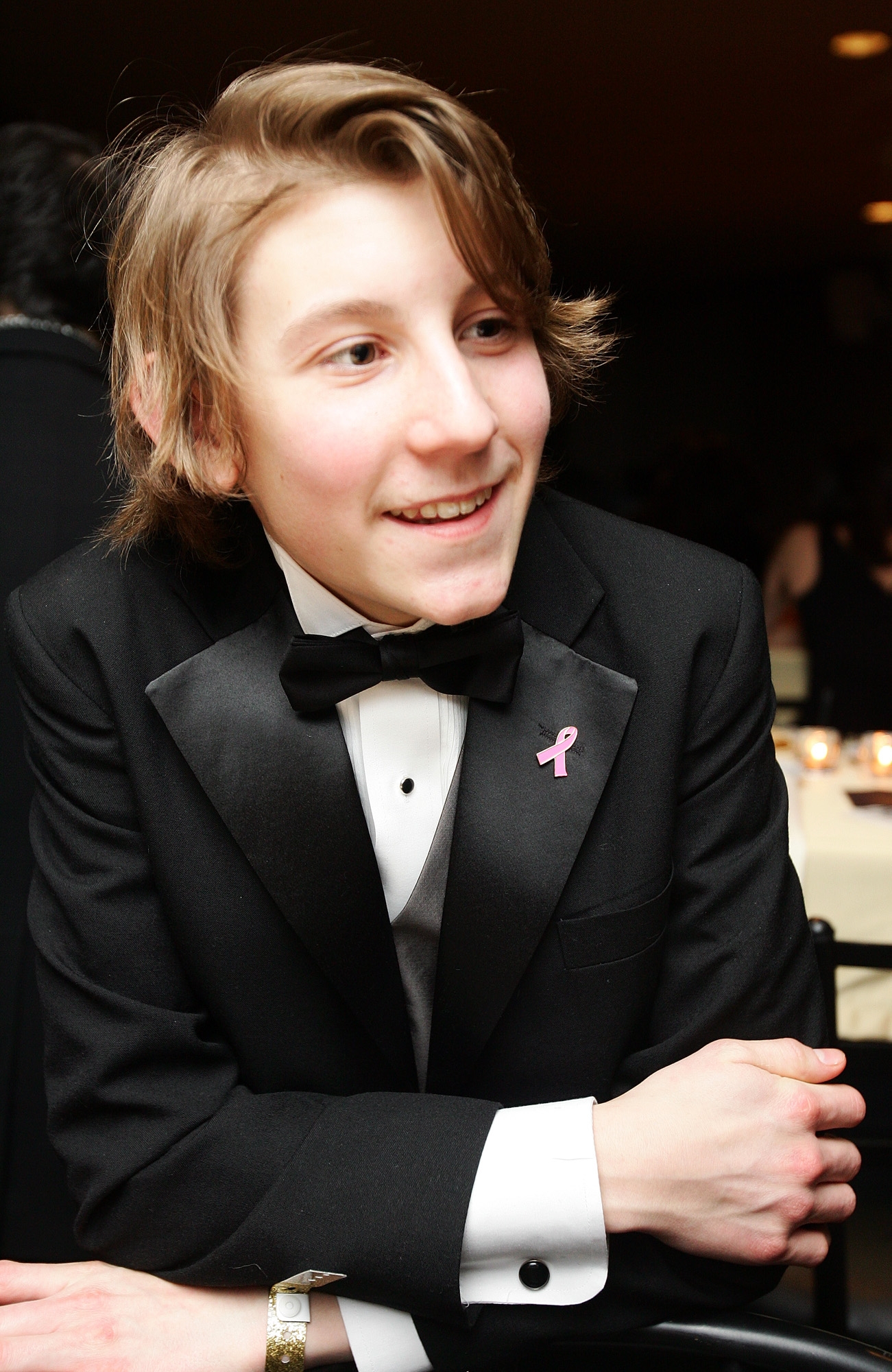 Teen in a tuxedo with a bow tie and a pink ribbon pin, smiling at an event