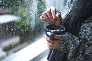 女性が雨の日に窓際でマグカップを持ち、ビスケットを浸している。リラックスした雰囲気。
