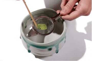 Hands sifting green matcha powder into a ceramic bowl