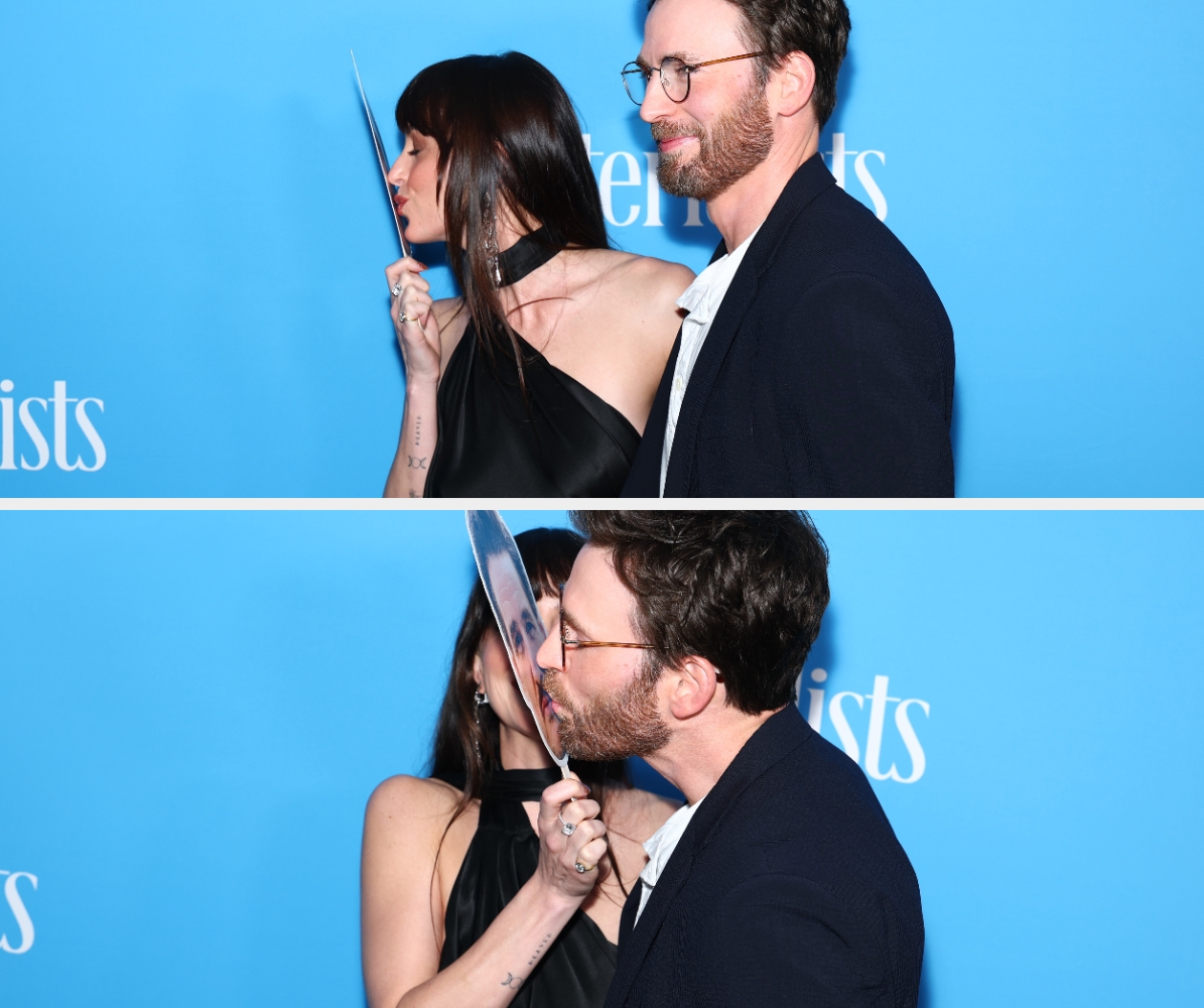 Two people on the red carpet, one in a black dress playfully holding a cutout. The other in a suit smiles beside them against a blue backdrop