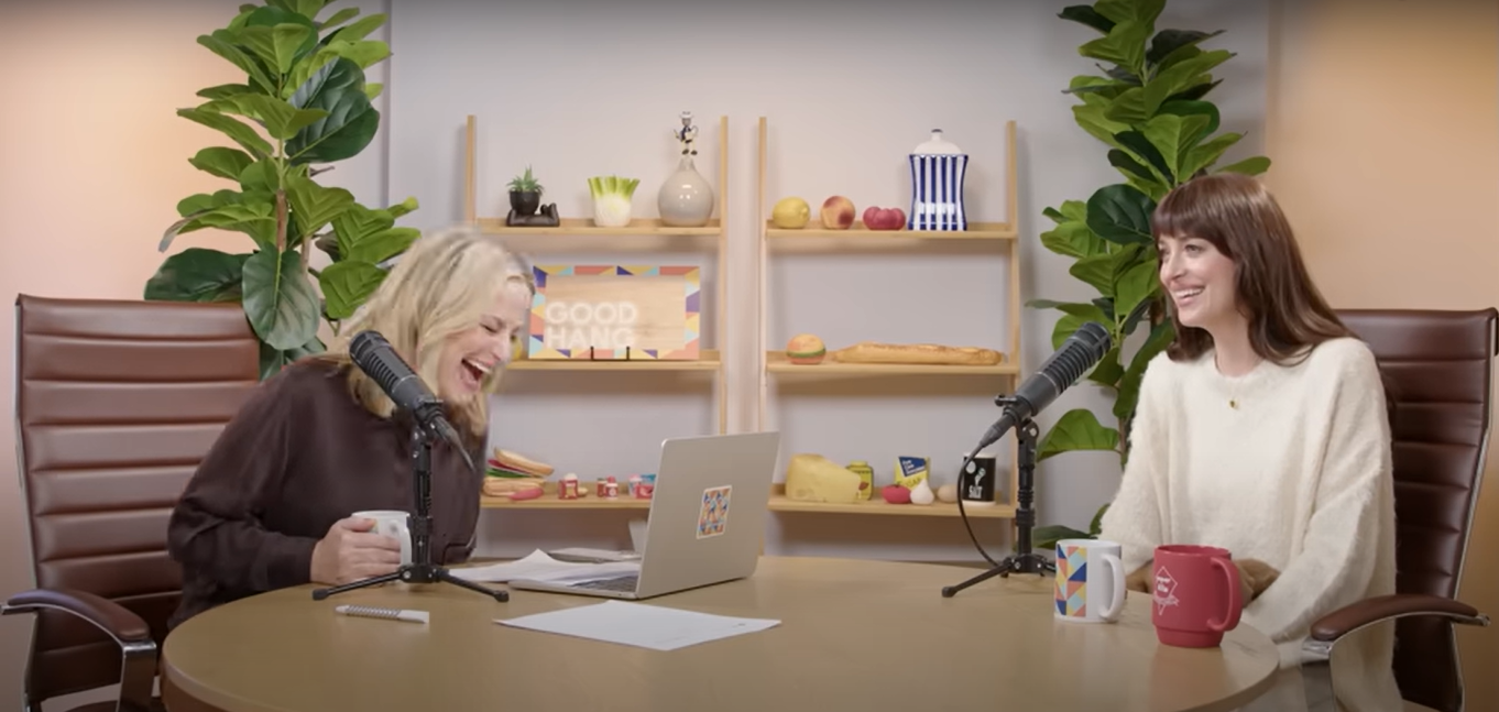 Two people laughing and talking into microphones during a podcast recording, seated at a table with mugs, papers, and plants in the background