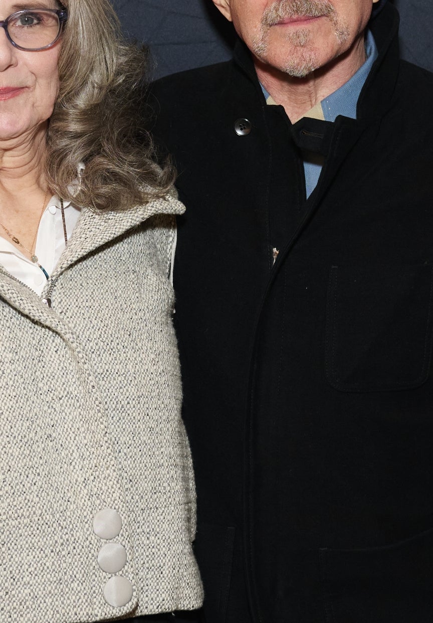 A man and woman stand together on a red carpet, each wearing glasses. The woman is in a casual outfit, while the man wears a dark suit