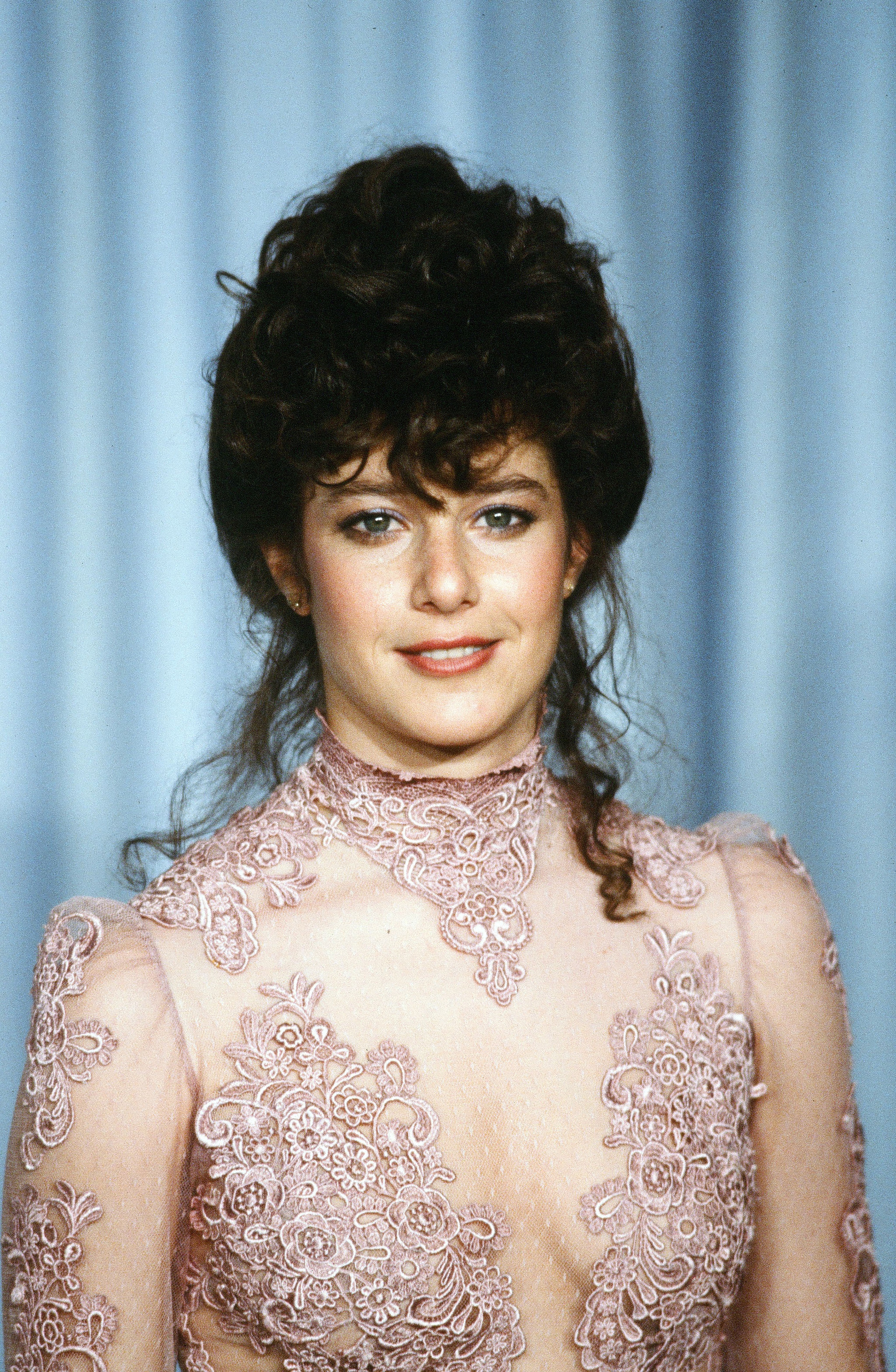 A person in a vintage lace dress with a high collar and intricate patterns stands in front of a curtain background