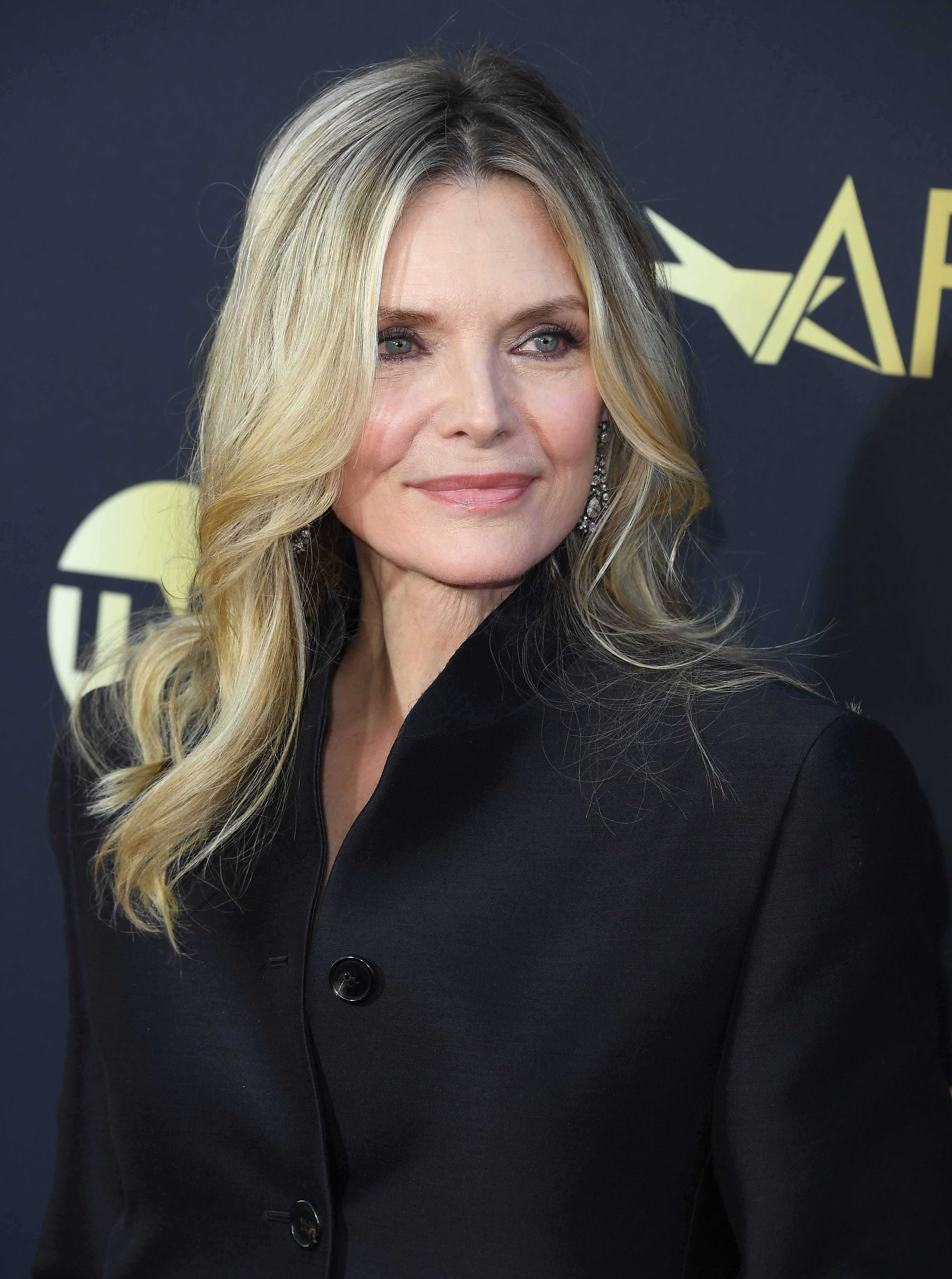 Person in a sleek black coat with long, wavy hair on a red carpet at a celebrity event