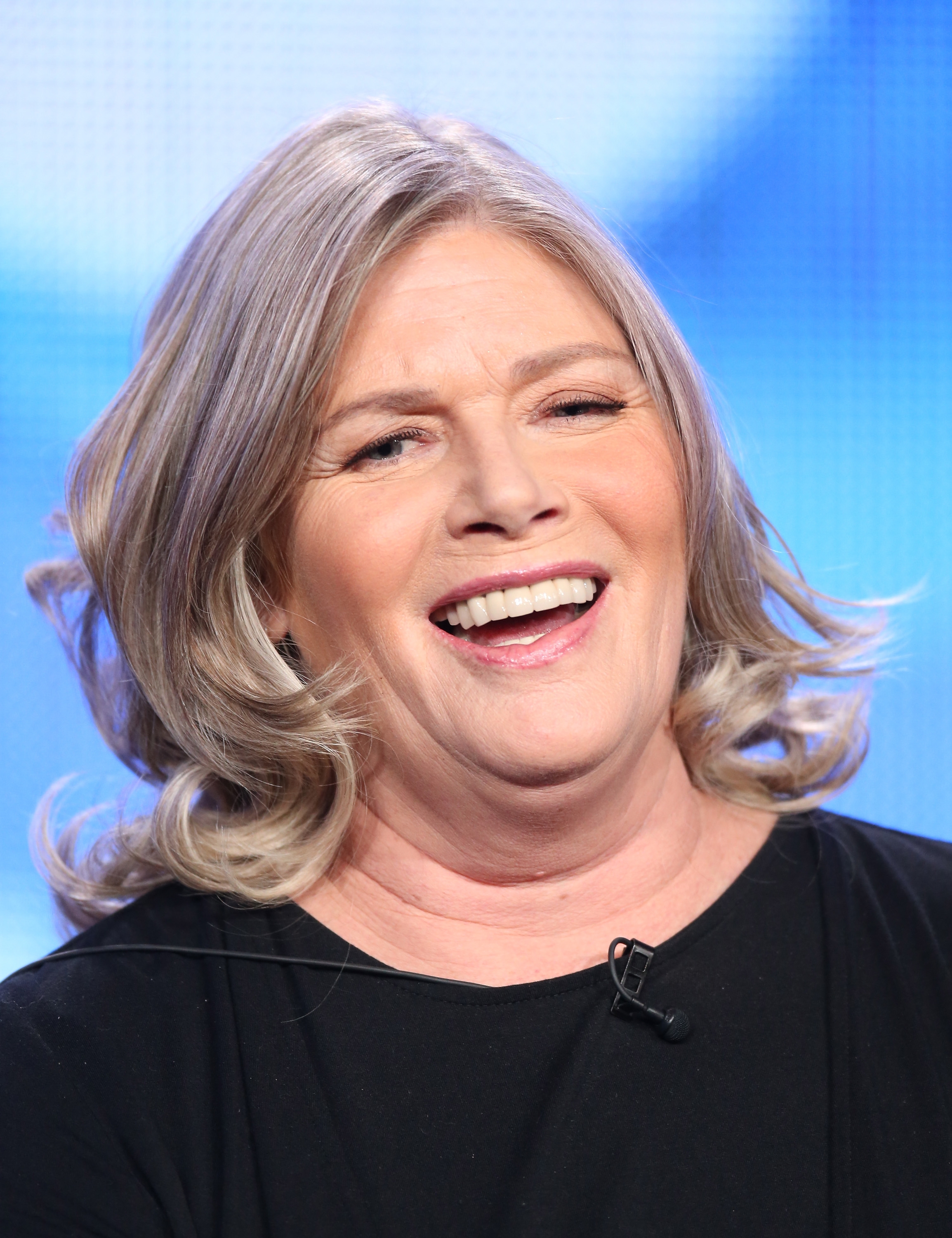 A person with shoulder-length hair smiles broadly, wearing a black top, possibly at a media event