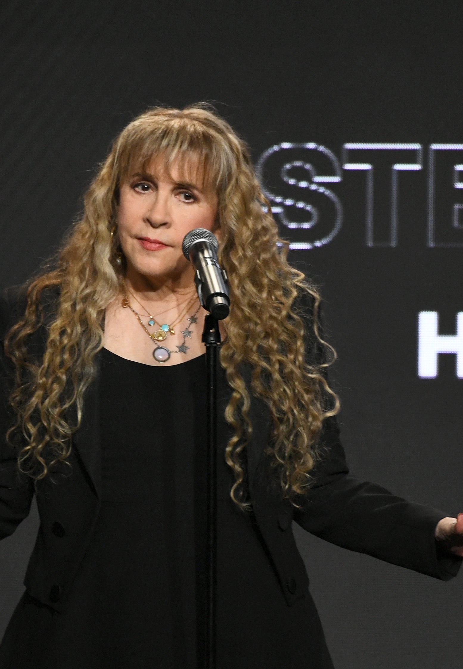 Person on stage with long wavy hair, wearing a black outfit, stands at microphone during an event. Text behind reads &quot;Hall of Fame.&quot;