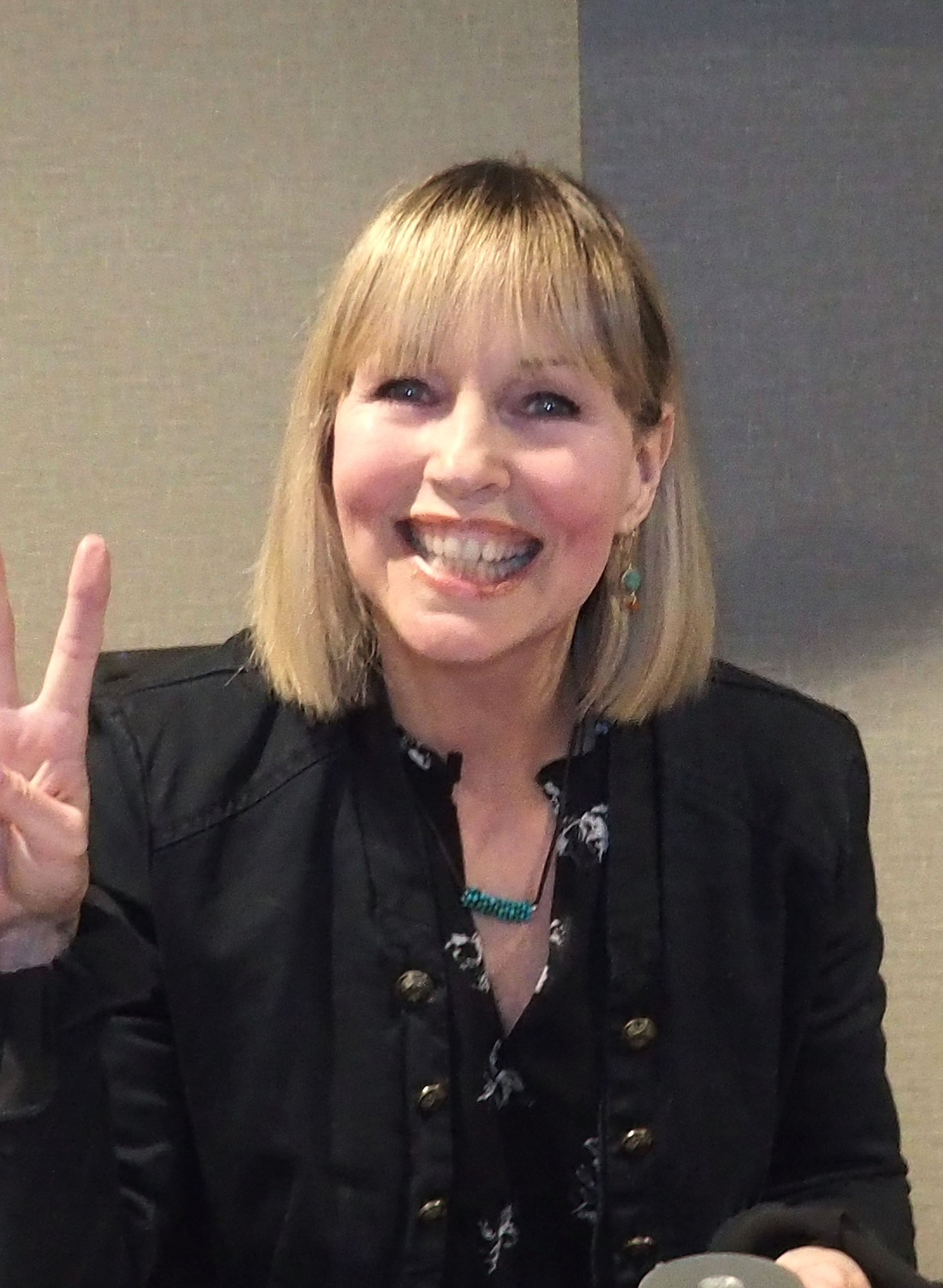 Person smiling, with shoulder-length hair, wearing a black jacket and patterned top, making a peace sign gesture