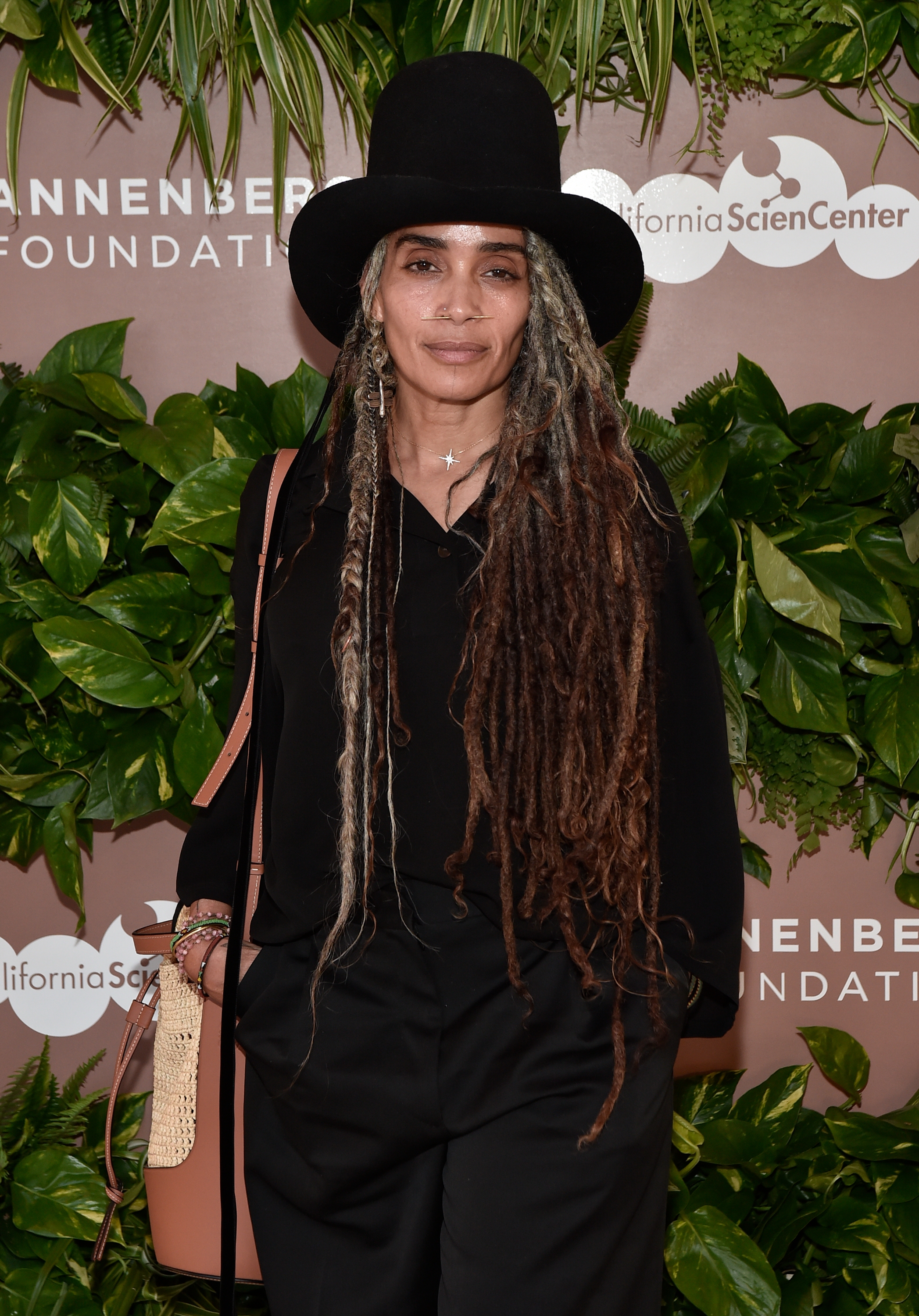 Person in a black outfit and large hat stands on a red carpet with a backdrop of green foliage