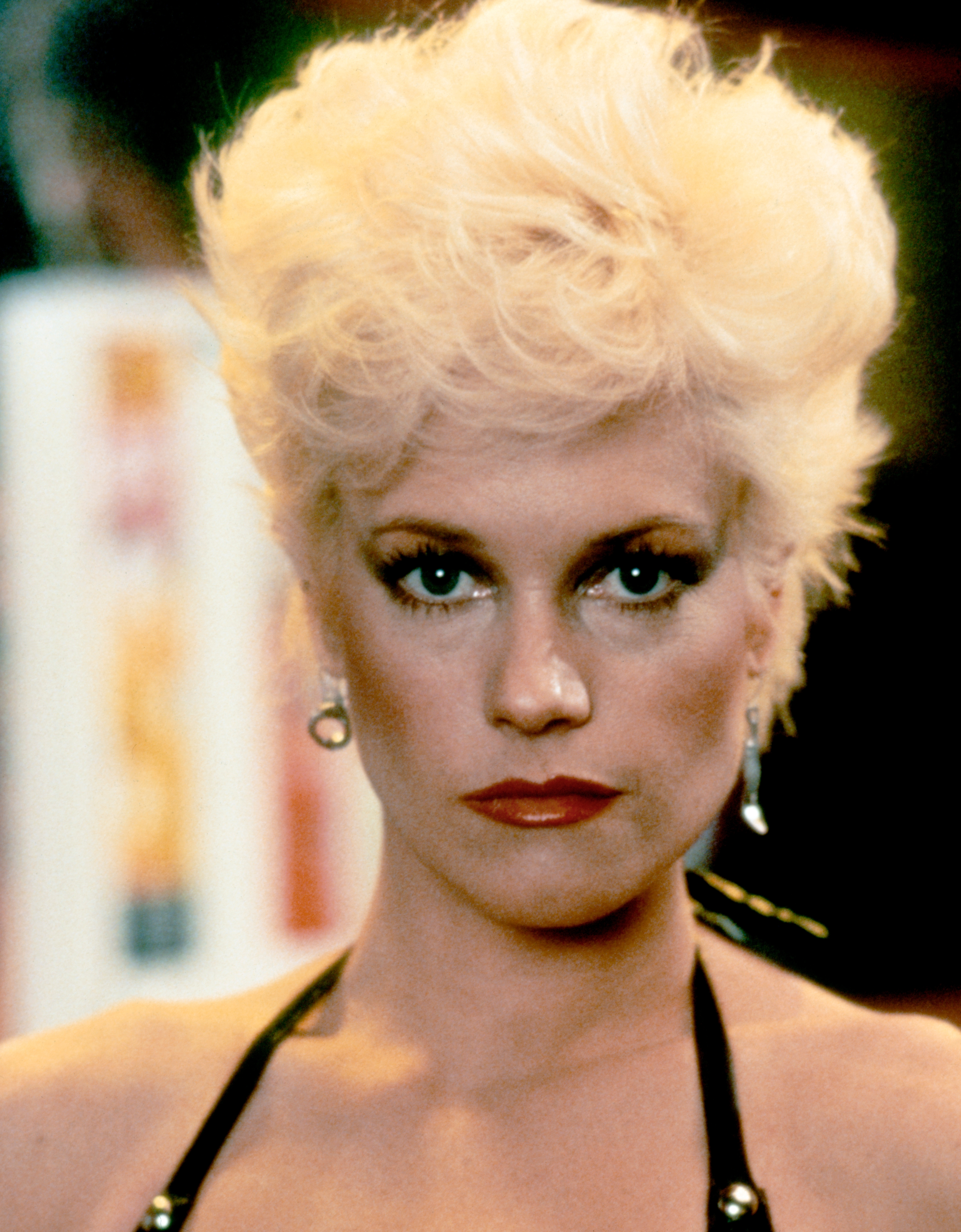 Person with short, voluminous hairstyle and bold makeup, wearing a halter top and hoop earrings, looking directly at the camera