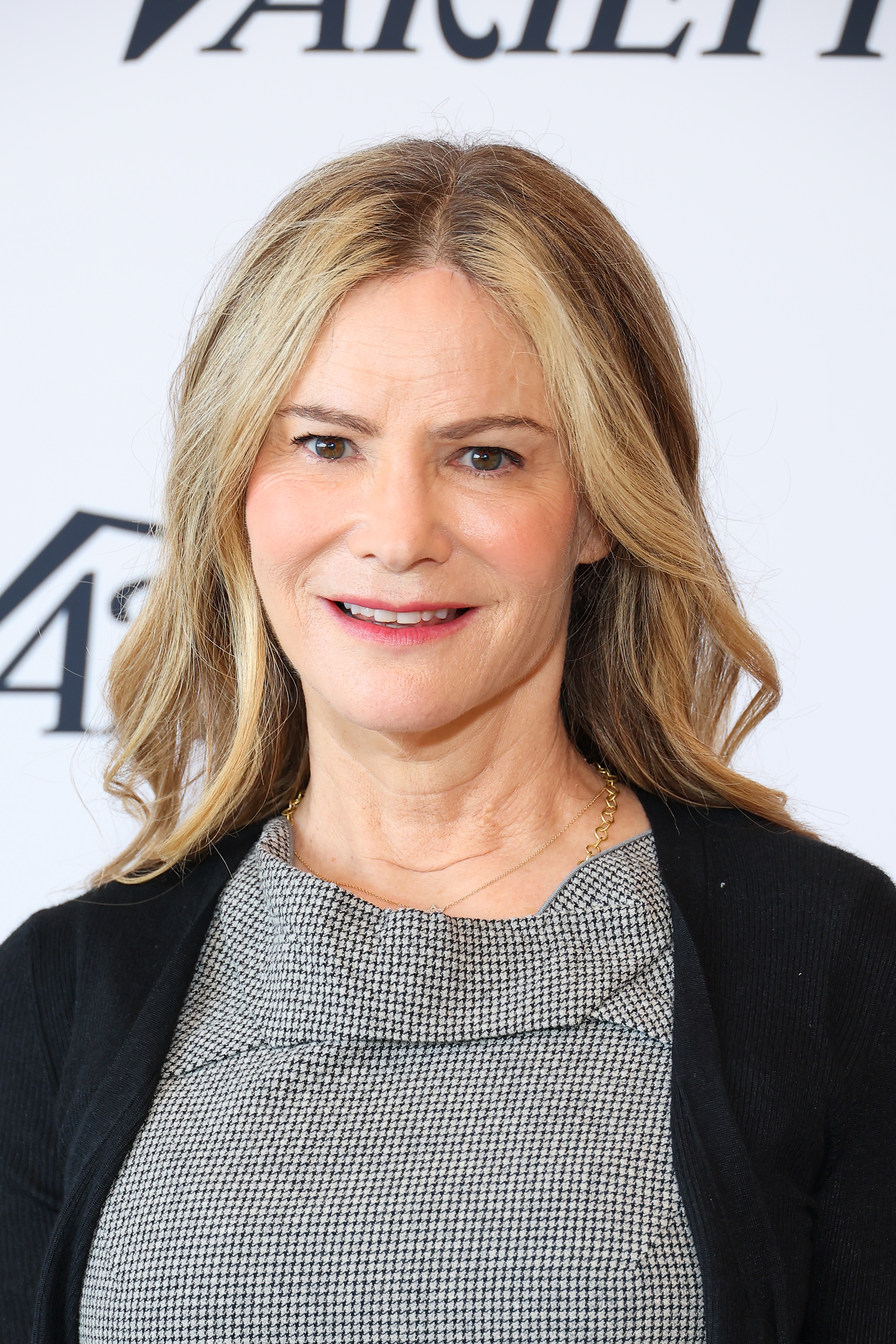 Person with long hair in a houndstooth dress and black cardigan, smiling at an event with a Variety backdrop