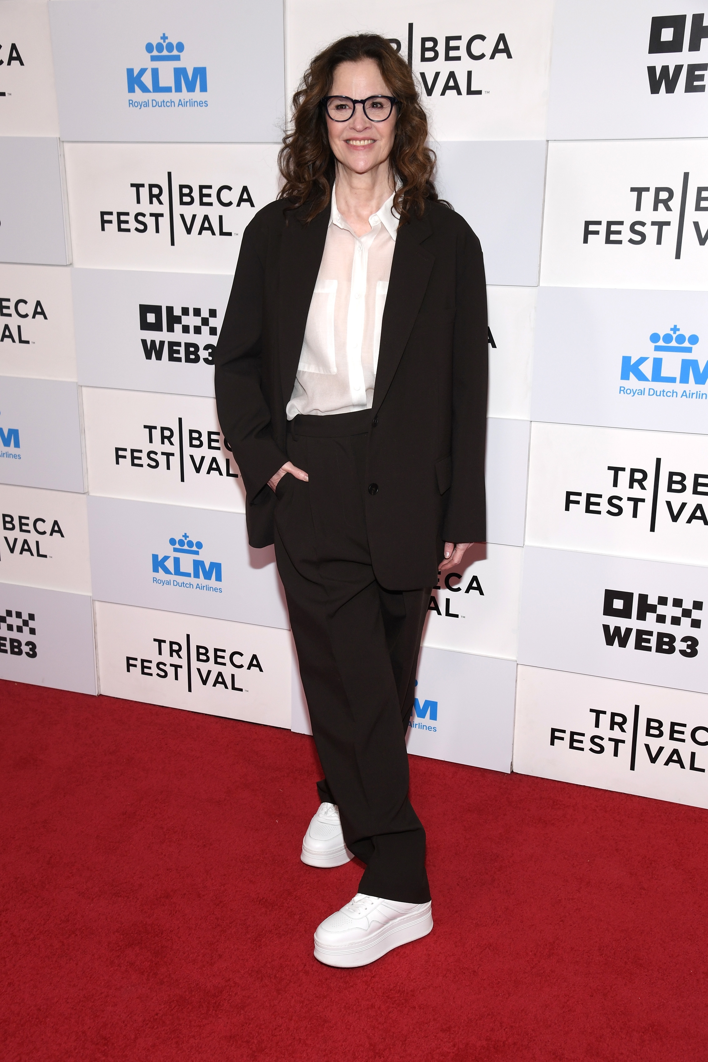 Person on red carpet in a black suit and white shirt, wearing glasses and white sneakers, standing in front of Tribeca Festival backdrop