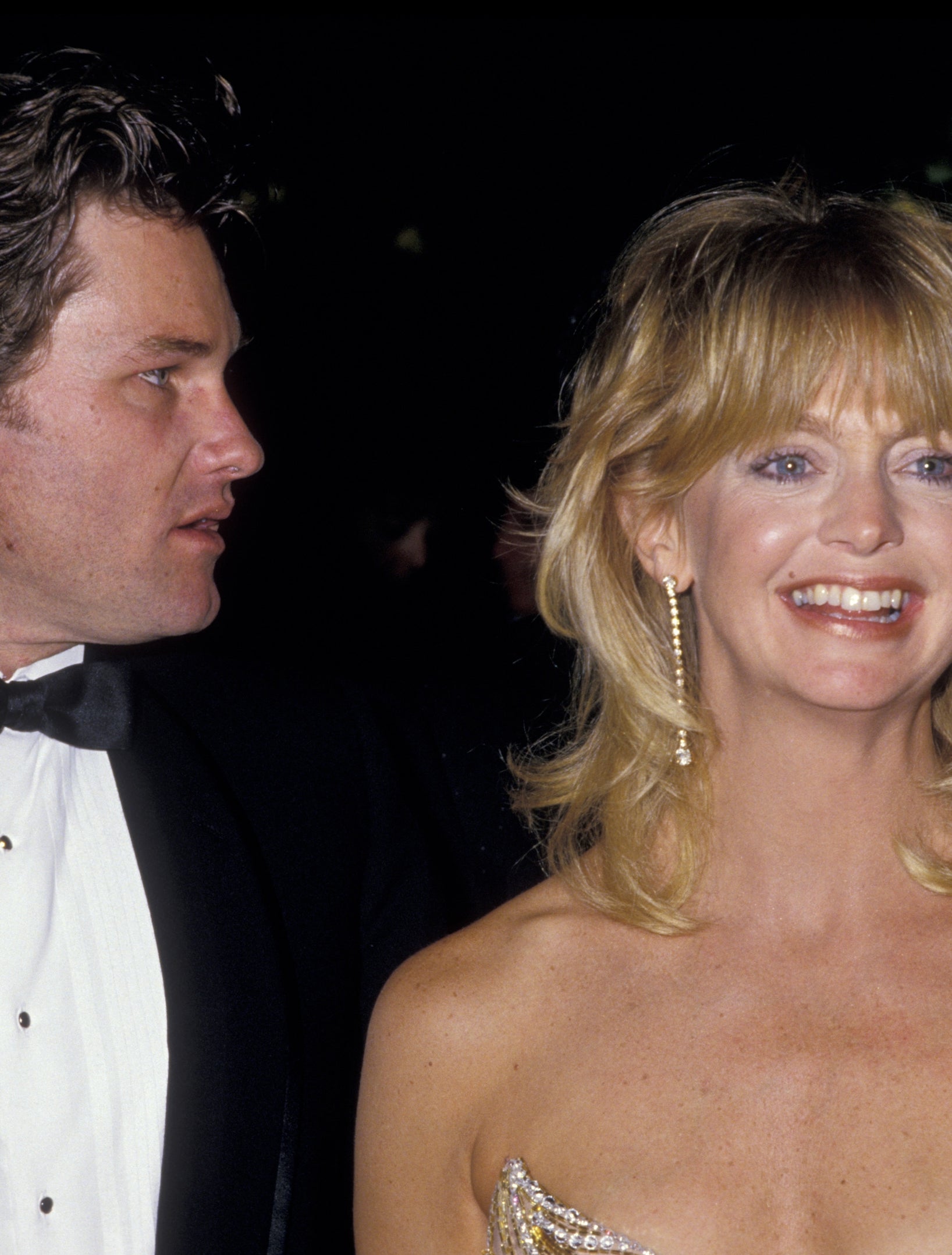 A couple poses on the red carpet. One wears a tuxedo, the other a strapless gown with gold details and large hoop earrings, both smiling