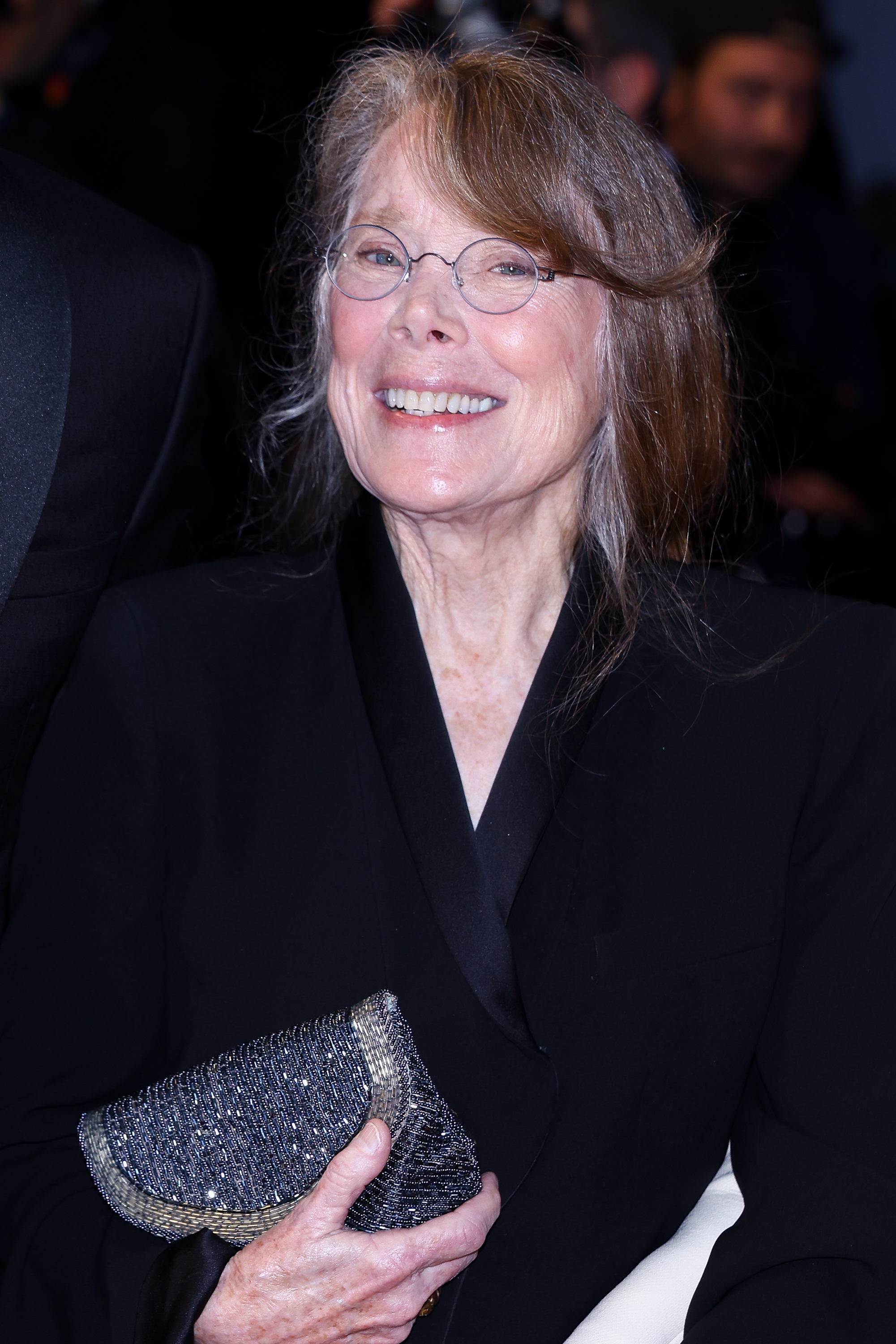 Person smiling at an event, wearing glasses and an elegant black outfit, holding a sparkling clutch purse