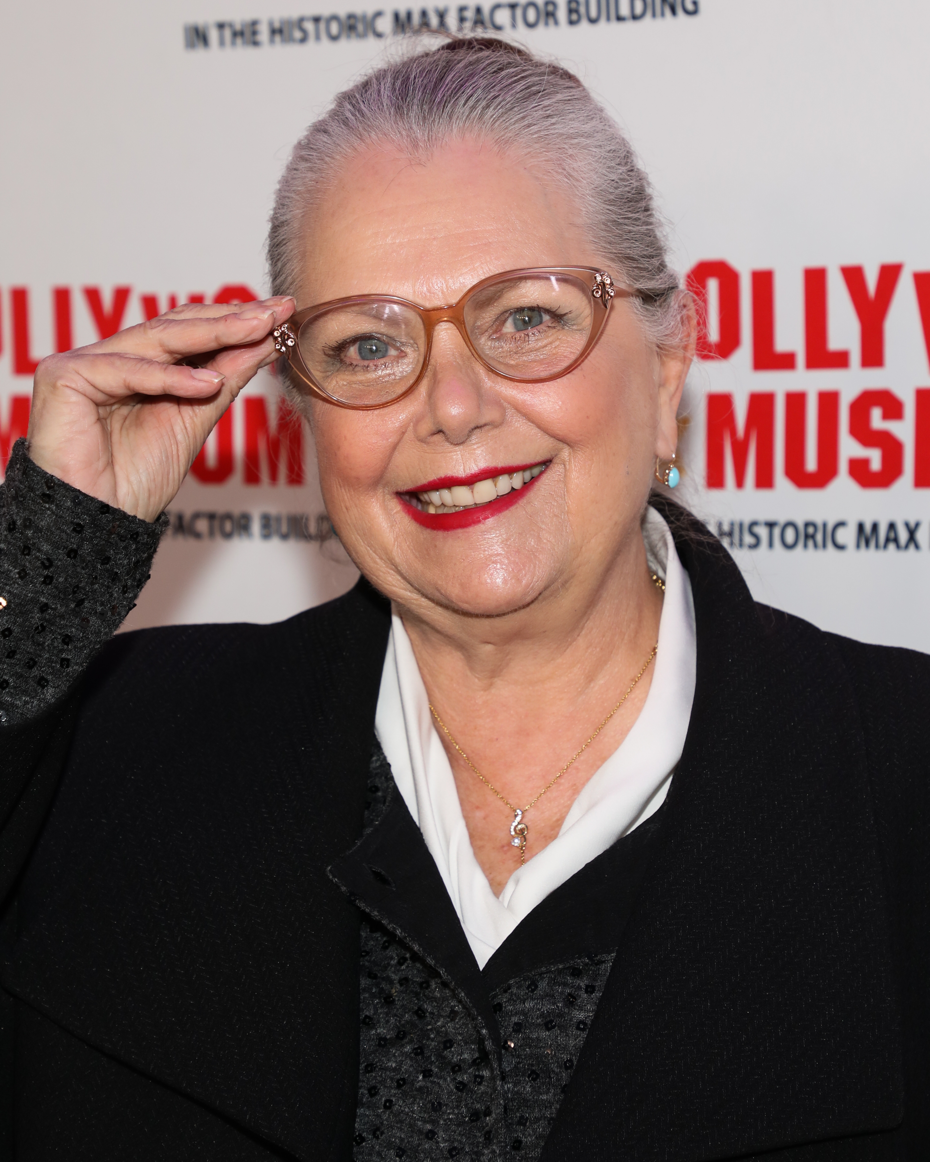 Person in elegant attire on the red carpet, smiling and adjusting stylish glasses. Background includes partial museum signage