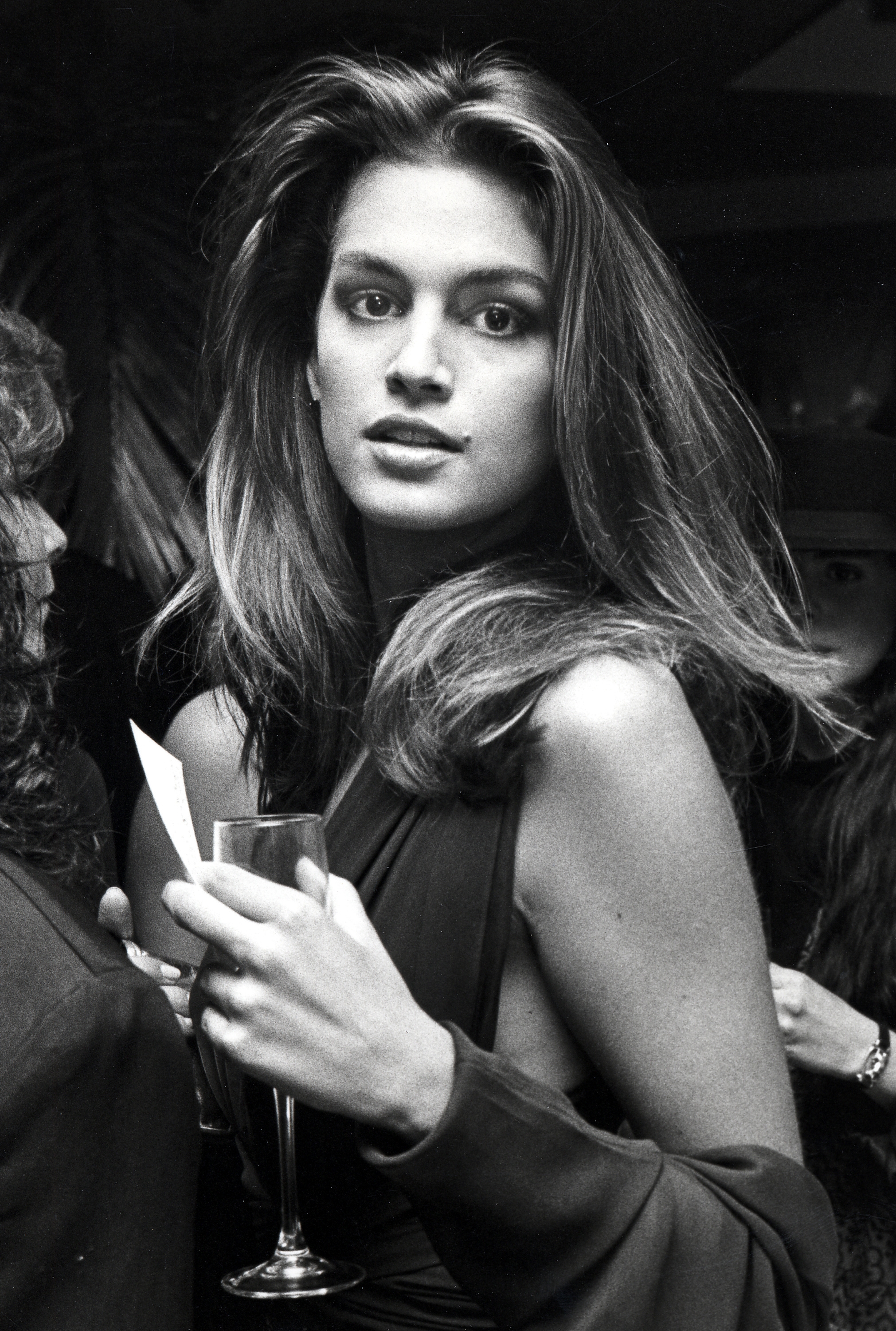 Person in elegant, sleeveless dress holding a champagne glass at an event, with notable hair volume and confident expression