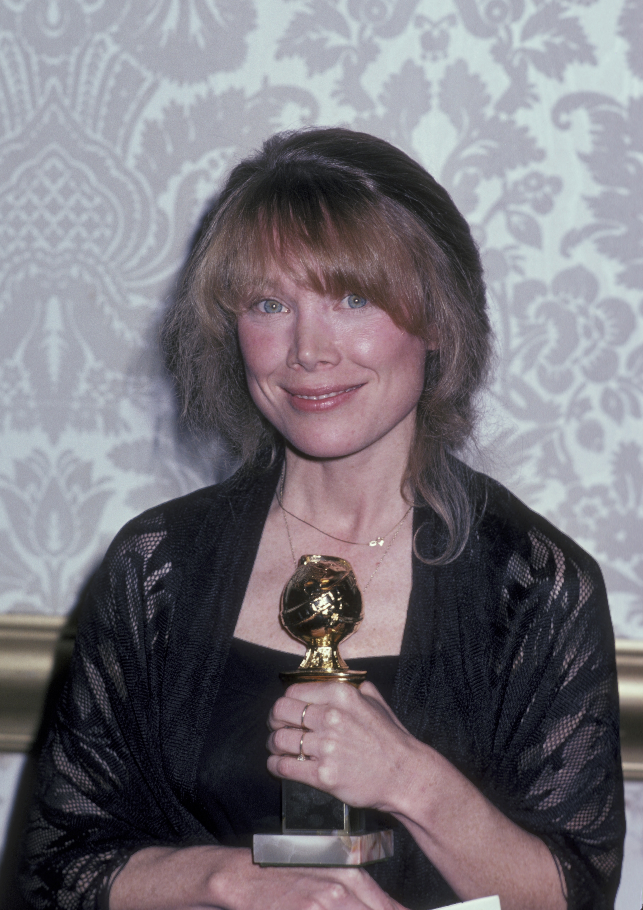 Person holding a Golden Globe award, wearing an elegant black dress with a delicate necklace, smiling at an event