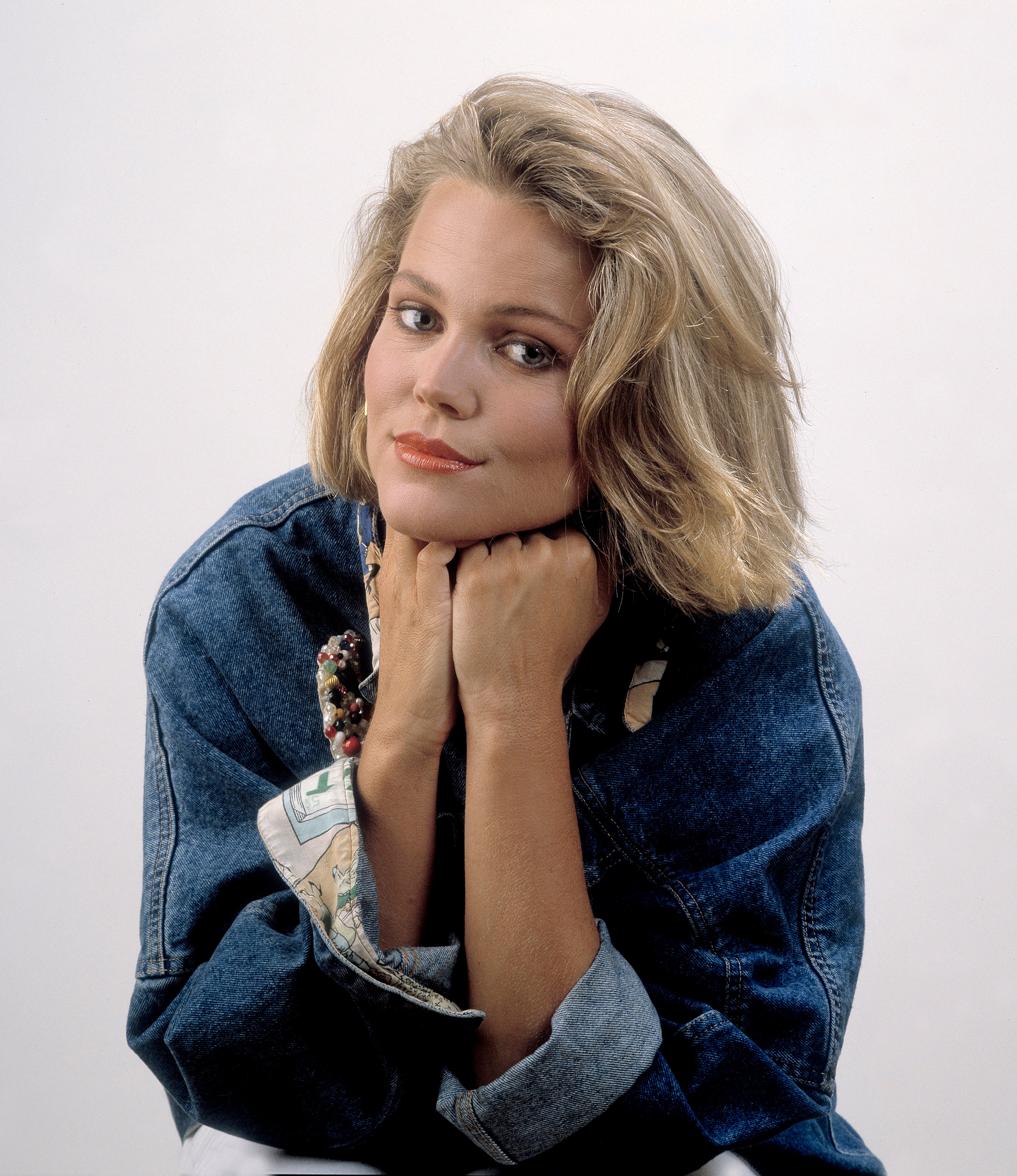 Person posing with head on hands, wearing a denim jacket and looking at the camera with a confident expression