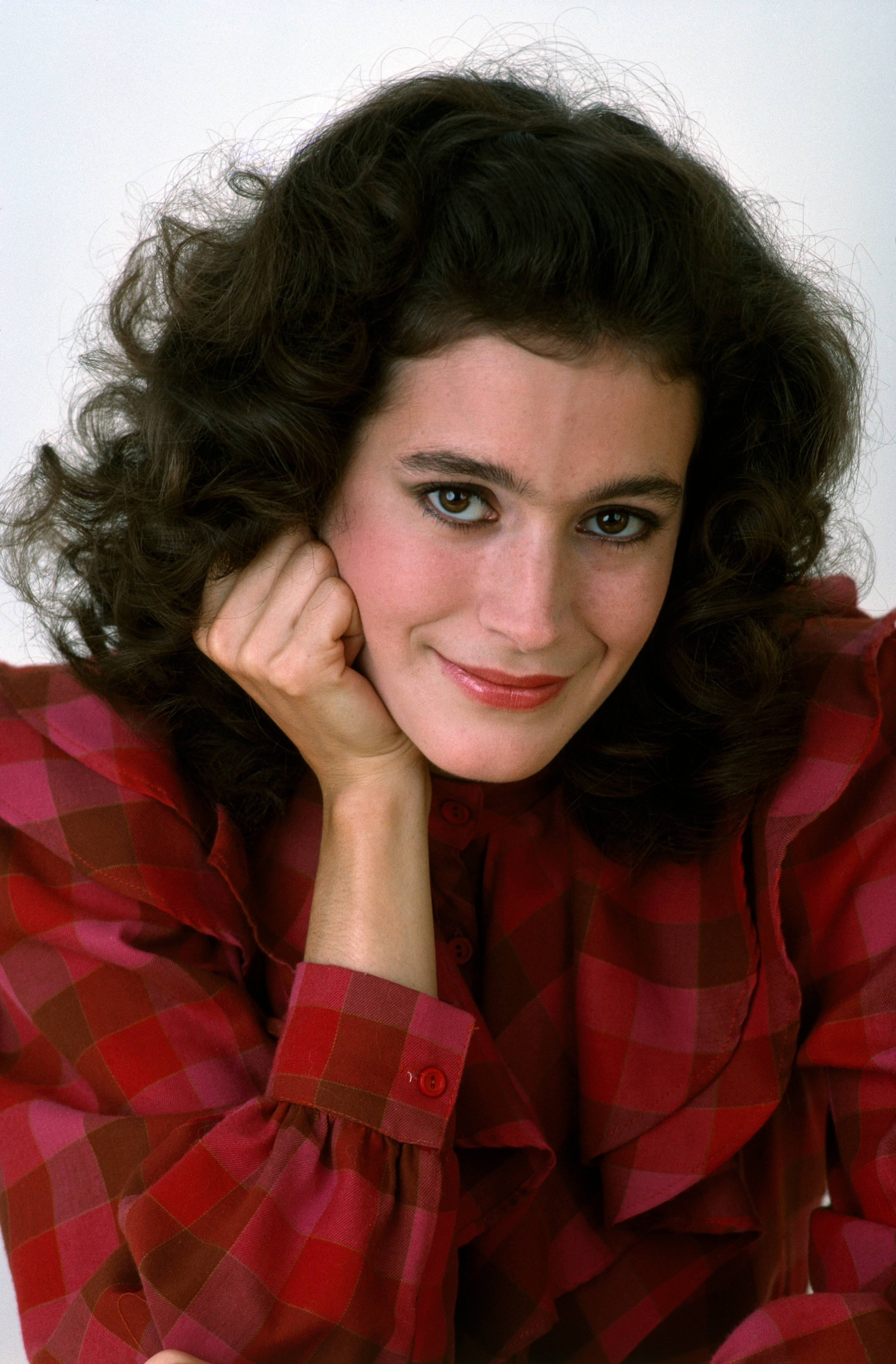 Person with curly hair poses with hand on cheek, wearing a checkered patterned blouse, smiling warmly at the camera
