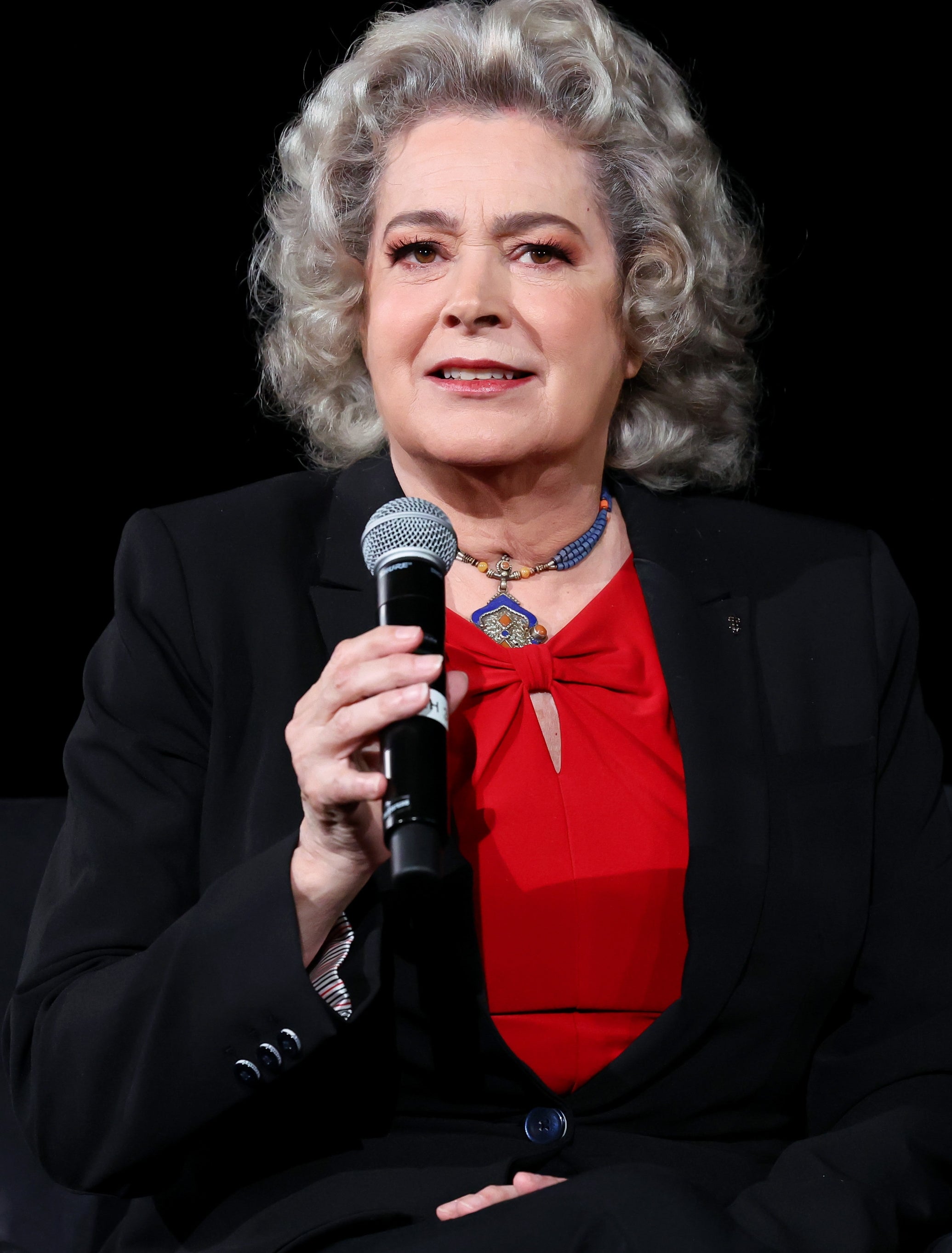 Person speaking into a microphone, wearing a dark suit with a bright top, sitting on a chair against a plain background