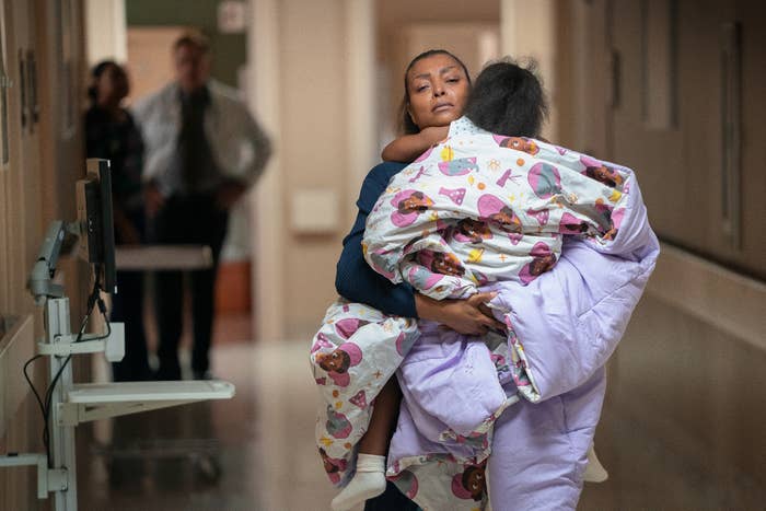 A woman looks concerned while carrying a child wrapped in a printed blanket down a hospital hallway