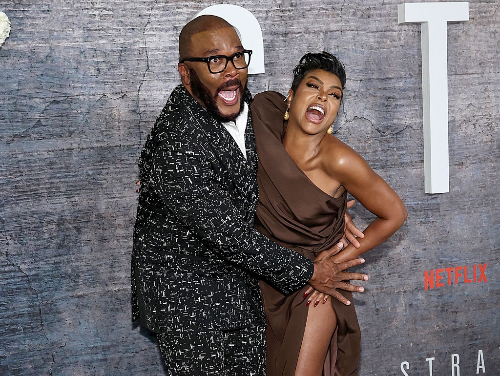Two people on the red carpet; one in a patterned suit, the other in a sleek, shoulder-baring gown, both laughing and playfully posing