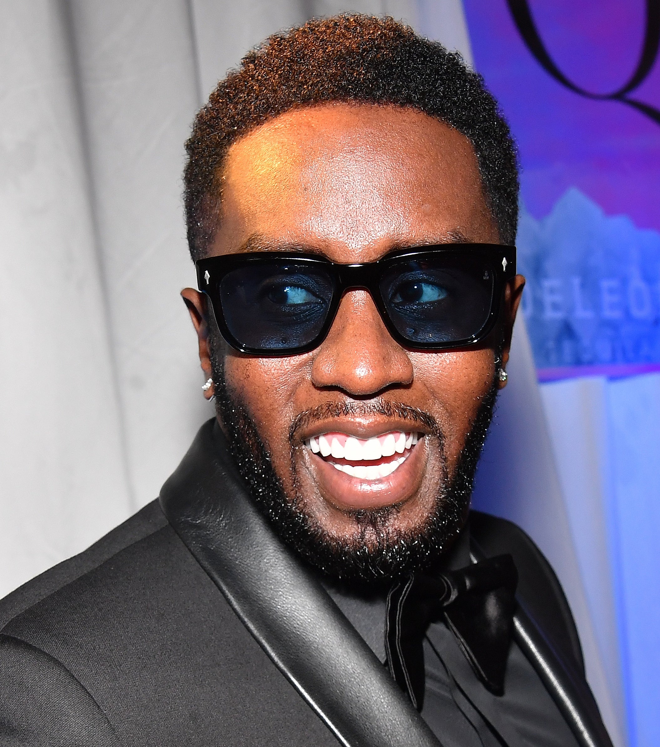 Diddy in a sleek black tuxedo and sunglasses, smiling at an event