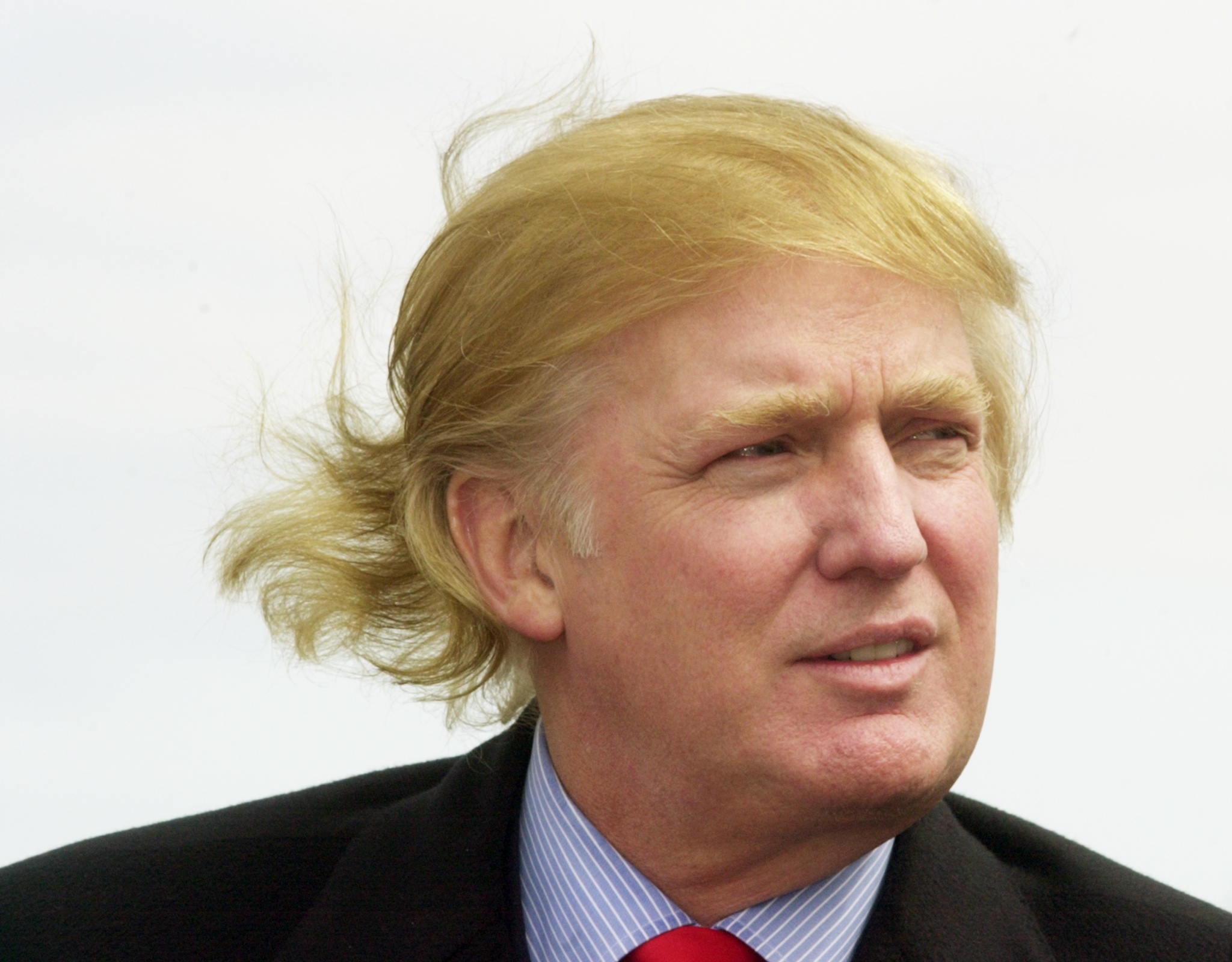 Trump with windblown hair looks to the side, wearing a suit with a striped shirt