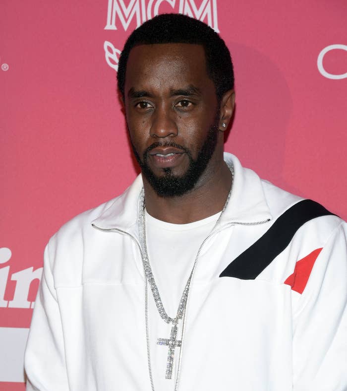 Sean "Diddy" Combs in a stylish white tracksuit with black accents and silver jewelry, standing against a backdrop of promotional logos