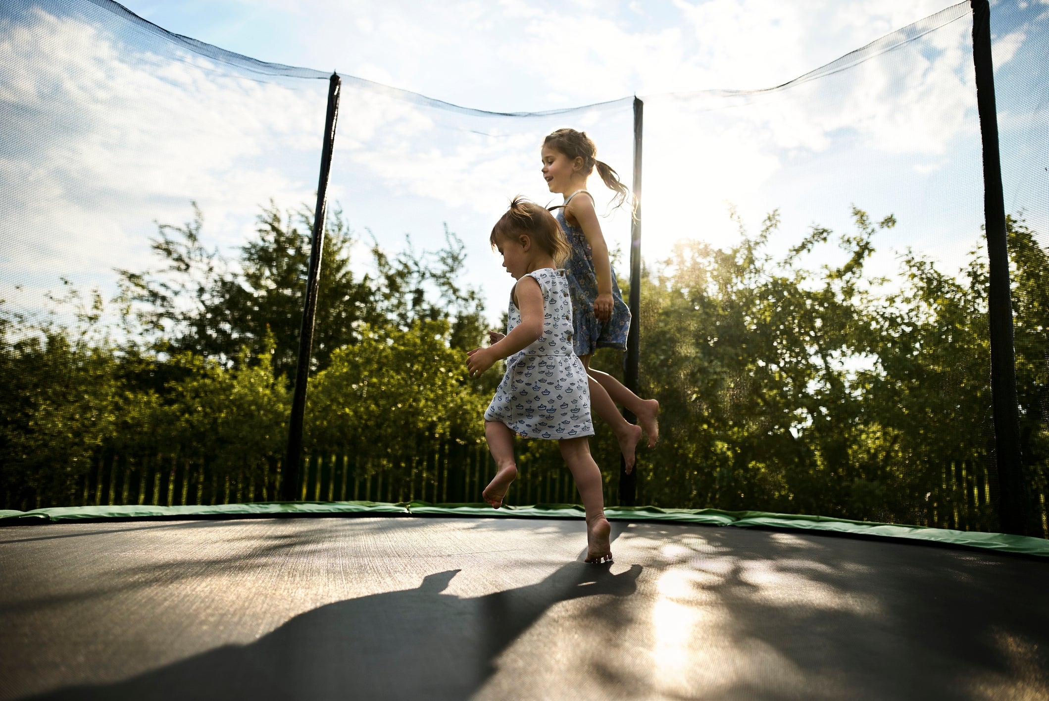 To små barn leker gledelig på en trampoline i en hage, med ett barn i luften og sollyset som strømmer gjennom trærne