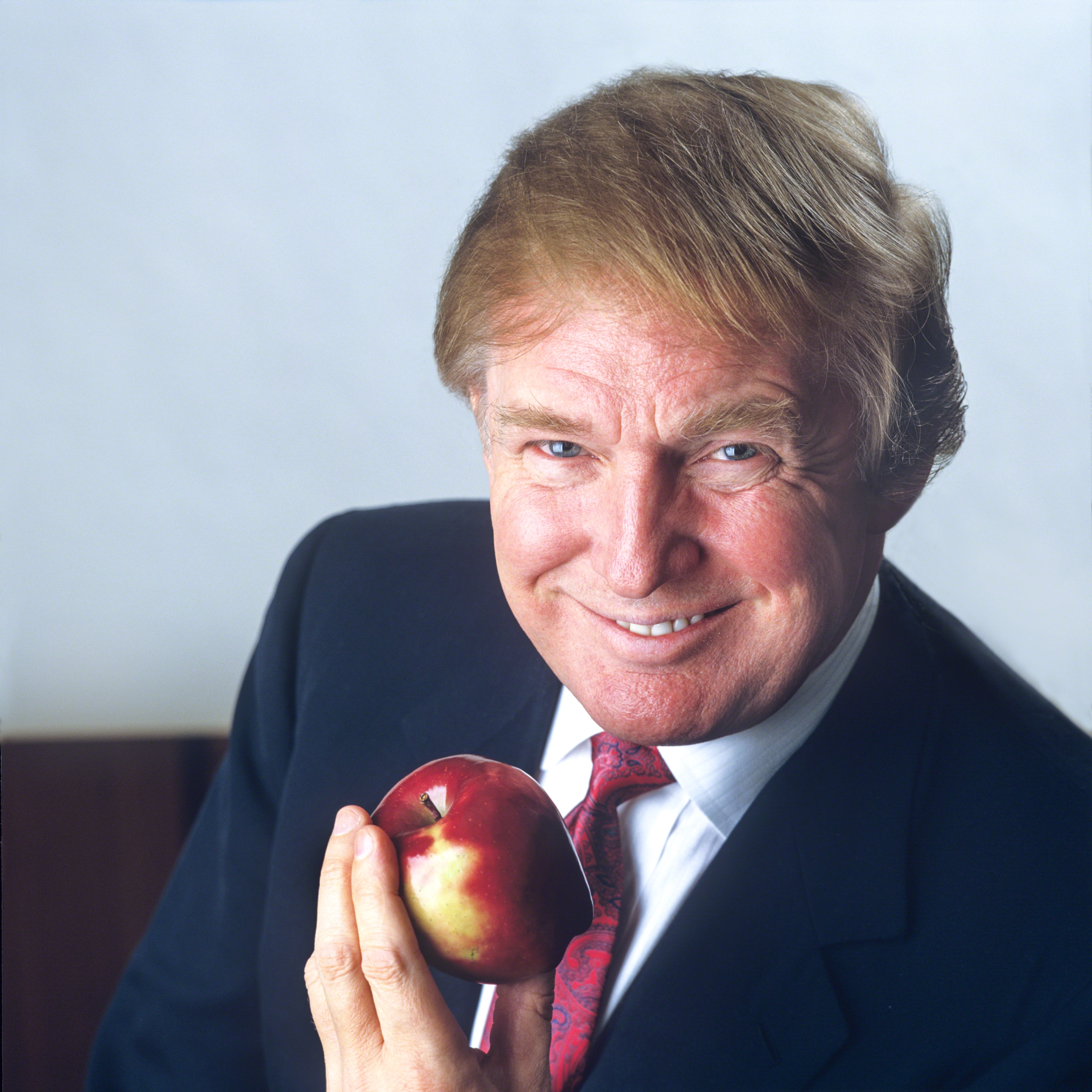 Trump holding an apple, smiling at the camera, wearing a suit and tie